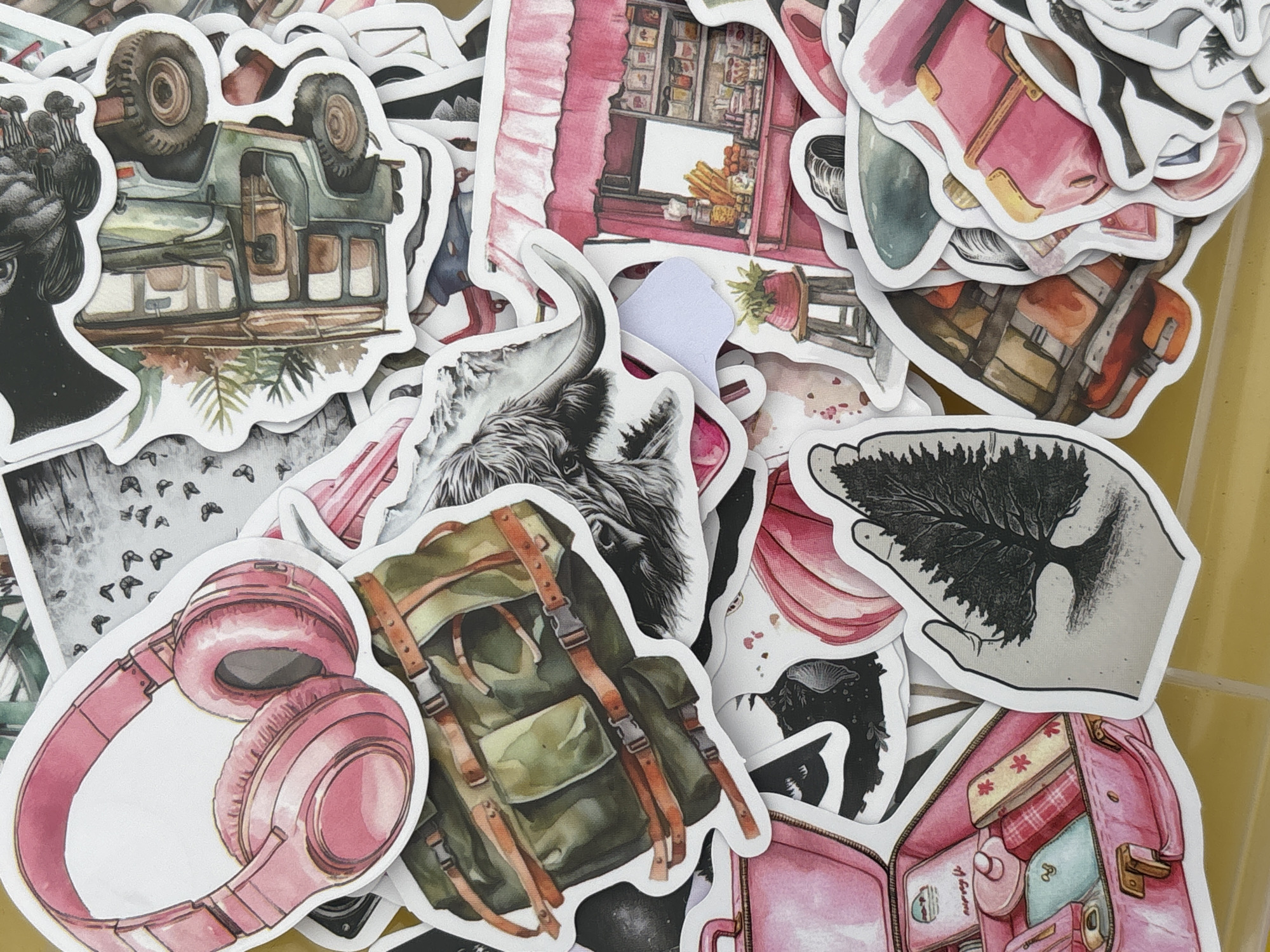 The image shows a pile of watercolor-style stickers. The stickers depict various subjects including: an overturned green off-road vehicle, a figure with mushrooms on its head, a pink storefront, a yak, pink headphones, a green backpack, a hand holding a tree-shaped black image, and a pink suitcase filled with items. The background is a yellowish-tan color. There are no people visible in the images on the stickers or in the background. There is no indication of the location Orenburg, Russia.
