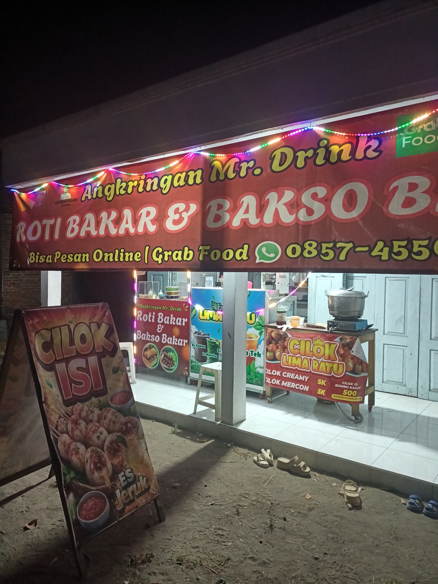 This is a night-time, eye-level, outdoor photograph of a food stall in Badas, Indonesia. The stall's banner reads "Angkringan Mr. Drink Roti Bakar & Bakso" and "Bisa Pesan Online | Grab Food 0857-4550". Colorful string lights are draped across the top of the banner. To the left of the stall, an A-frame sign displays images of "CILOK ISI" and "ES Jeruk." Inside the stall, a pot sits on a portable gas stove. Another sign on the stall advertises "CILOK LIMA RATU" with prices for "CILOK CREAMY," "CILOK MERCON," and "CILOK ISI BUMBU KACANG." Several pairs of sandals are scattered on the sandy ground in front of the stall. The lighting is dim, with the stall and its signs being the primary illuminated objects.