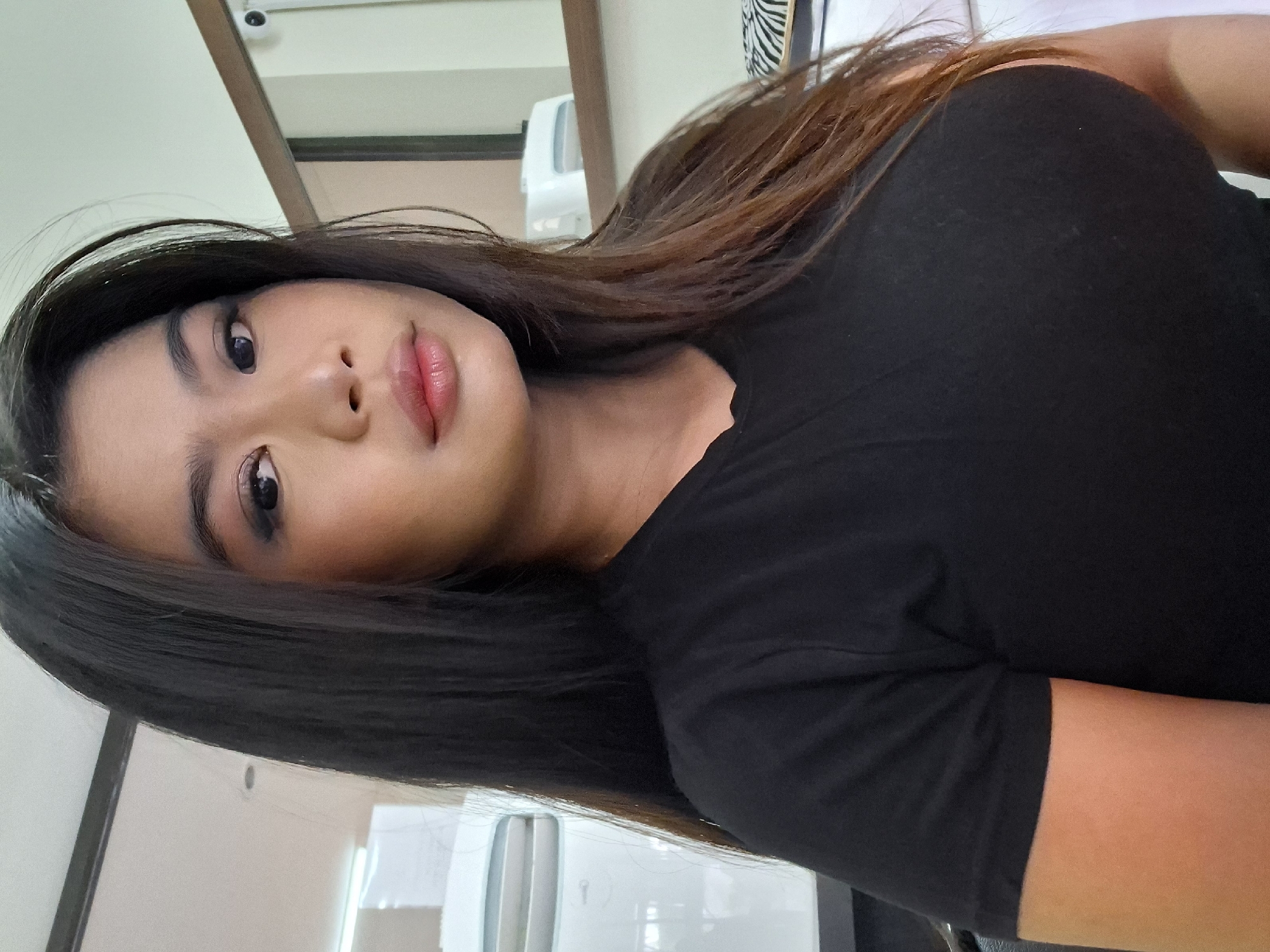 A woman with long dark hair is facing the camera, her head angled slightly to her left. She is wearing a black t-shirt. Her makeup includes dark eyeliner and lipstick. Behind her, on the left, is a white appliance, possibly a refrigerator. To the right, a mirror reflects a shelf with a white object. The setting appears to be indoors, possibly in a residential or office space. There is no visible indication of Kuta, Indonesia.