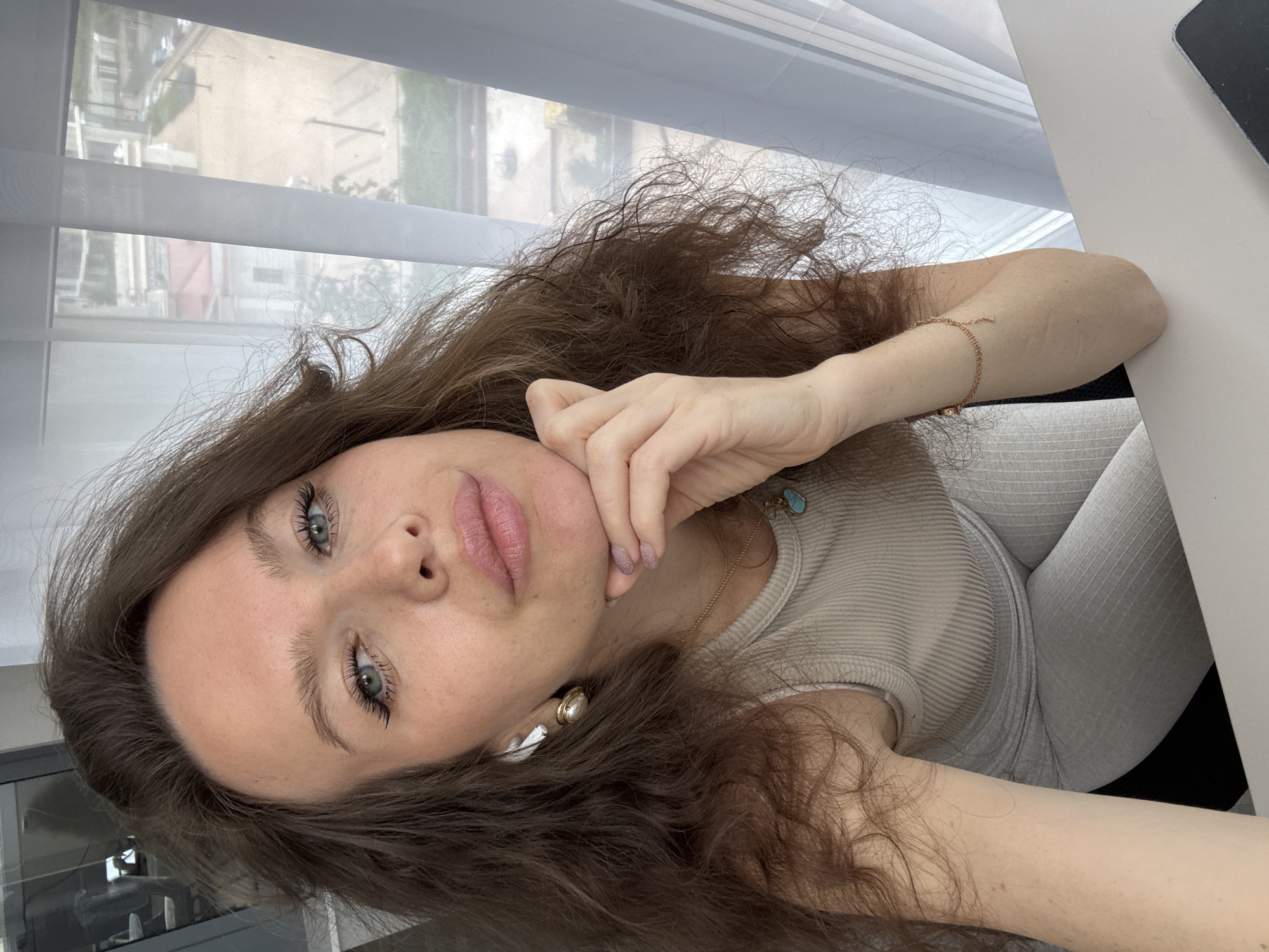 A close-up, tilted portrait captures a young woman indoors in Khimki, Russia. She has long, wavy brown hair, striking blue-grey eyes, and appears to be looking directly at the camera. Her expression is neutral to slightly pensive, with her light pink lips slightly parted. Her right hand rests gently against her chin, and her fingernails have a faint, glittery polish.
She is wearing a light beige or tan ribbed tank top and light grey ribbed leggings or pants. Accessories include small golden hoop earrings with pearl-like drops, white wireless earbuds, a delicate gold chain necklace with a small turquoise pendant, and a thin gold bracelet on her right wrist.
The setting appears to be a room with a large window in the background, featuring white horizontal blinds. Natural daylight streams in through the window, indicating it is daytime. Outside the window, an urban landscape is visible, partially obscured by the blinds. Buildings with multiple stories, some with visible windows and hints of green foliage, suggest a residential or mixed-use area. A light-colored table or desk is visible on the right side of the frame, with a dark object, possibly a smartphone, resting on it. No visible text is present in the image. The overall impression is a casual, self-taken portrait.