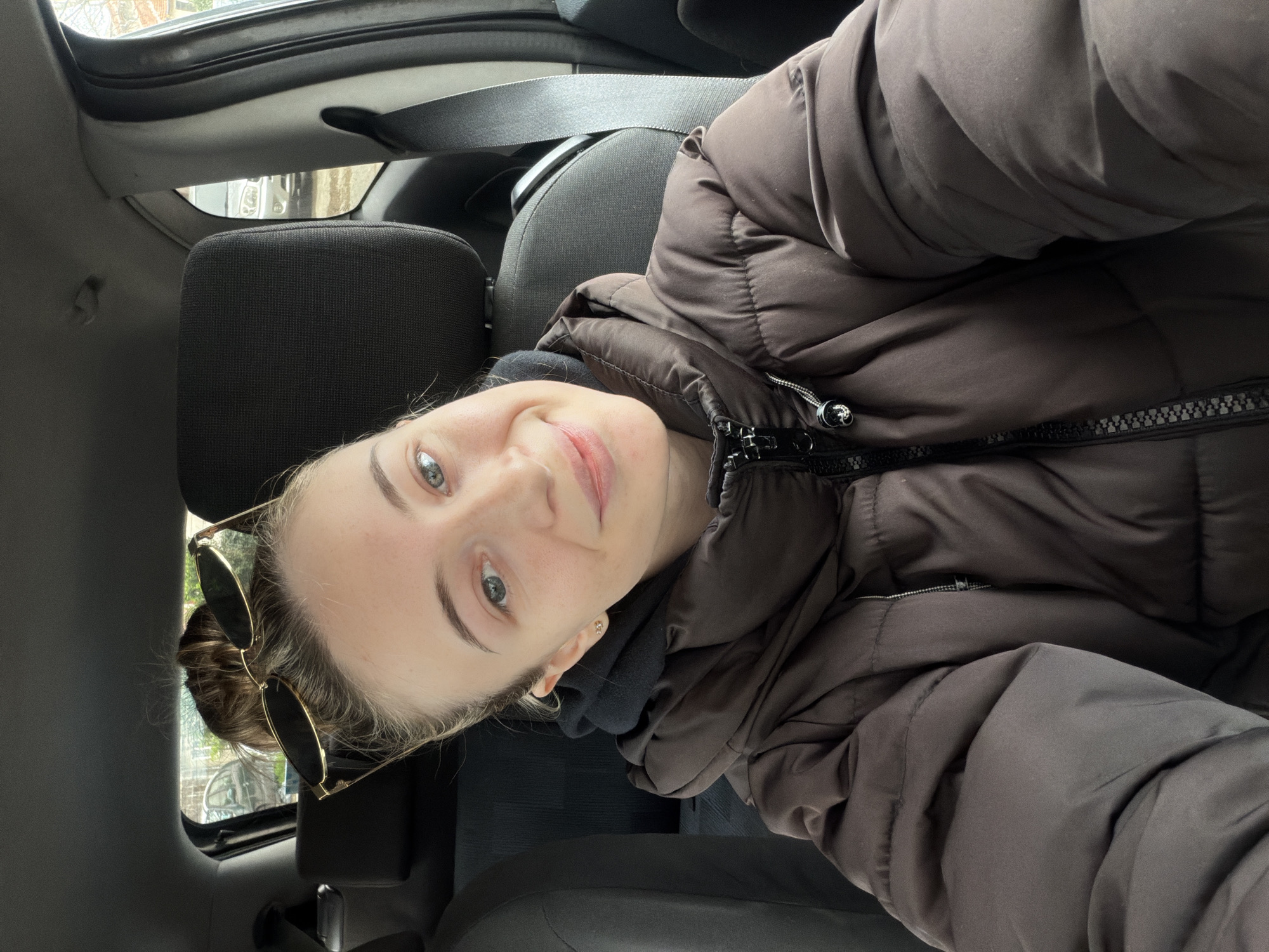 A person is in a car, likely a selfie based on the angle. The person is wearing a brown puffer jacket over a black hoodie and has sunglasses perched on their head. A seatbelt is visible in the background, along with the car's interior. Based on the information provided, no details in the image suggest this scene is in Plovdiv, Bulgaria. More information is needed to determine the specific location.
