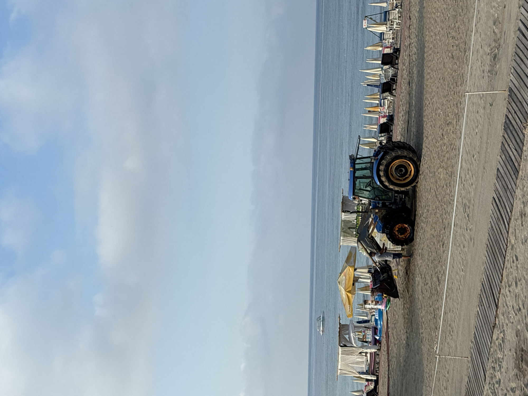 A blue tractor with a front loader is parked on a sandy beach in Alanya, Turkey, near the Mediterranean Sea. The tractor's bucket is lowered. A person in a light-colored t-shirt and dark shorts stands between the tractor and a shallow inflatable pool, appearing to be in the process of cleaning or organizing the area. In the background, rows of white beach umbrellas and loungers are set up along the coastline. The sea is calm and blue, with a white boat visible in the distance. The sky is partly cloudy with a light blue hue. A wooden pathway is visible in the foreground of the image.