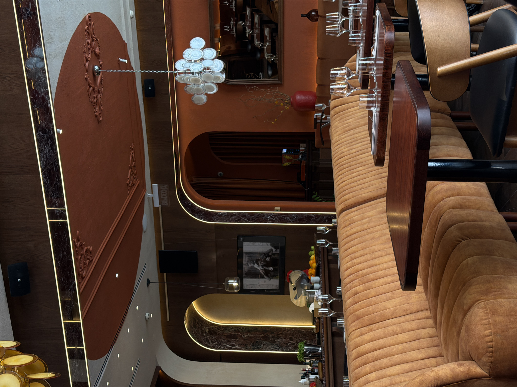 The image shows an interior view of a restaurant in Saint Petersburg, Russia. The scene is set in a dimly lit dining area, characterized by warm, earthy tones and elegant decor. In the foreground, a plush, ribbed, caramel-colored banquette curves around a dark wooden table, set with glassware and cutlery. Another similar table and banquette are visible further back.
The walls are adorned with arched niches, one of which houses a television screen displaying a black and white image. Overhead, a decorative ceiling features intricate molding and a modern chandelier composed of translucent discs. A sign with Russian text, likely indicating an exit ("ВЫХОД"), is visible on the wall. The overall ambiance suggests a sophisticated and contemporary dining establishment.