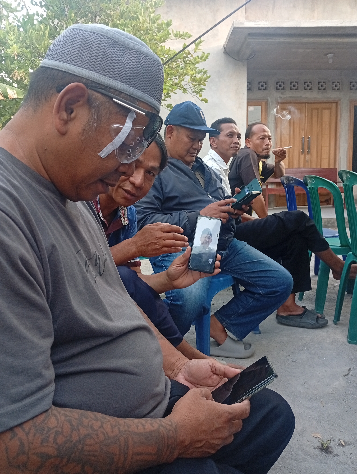 A group of five middle-aged Indonesian men are seated outdoors, appearing to be in the city of Delanggu, Indonesia. The men are dressed casually, with some wearing traditional Indonesian caps. Two of the men are holding and looking at their mobile phones. One man is smoking a cigarette. The environment appears to be a courtyard or open area with a building in the background, and plastic chairs are arranged for seating. The lighting suggests it is daytime.