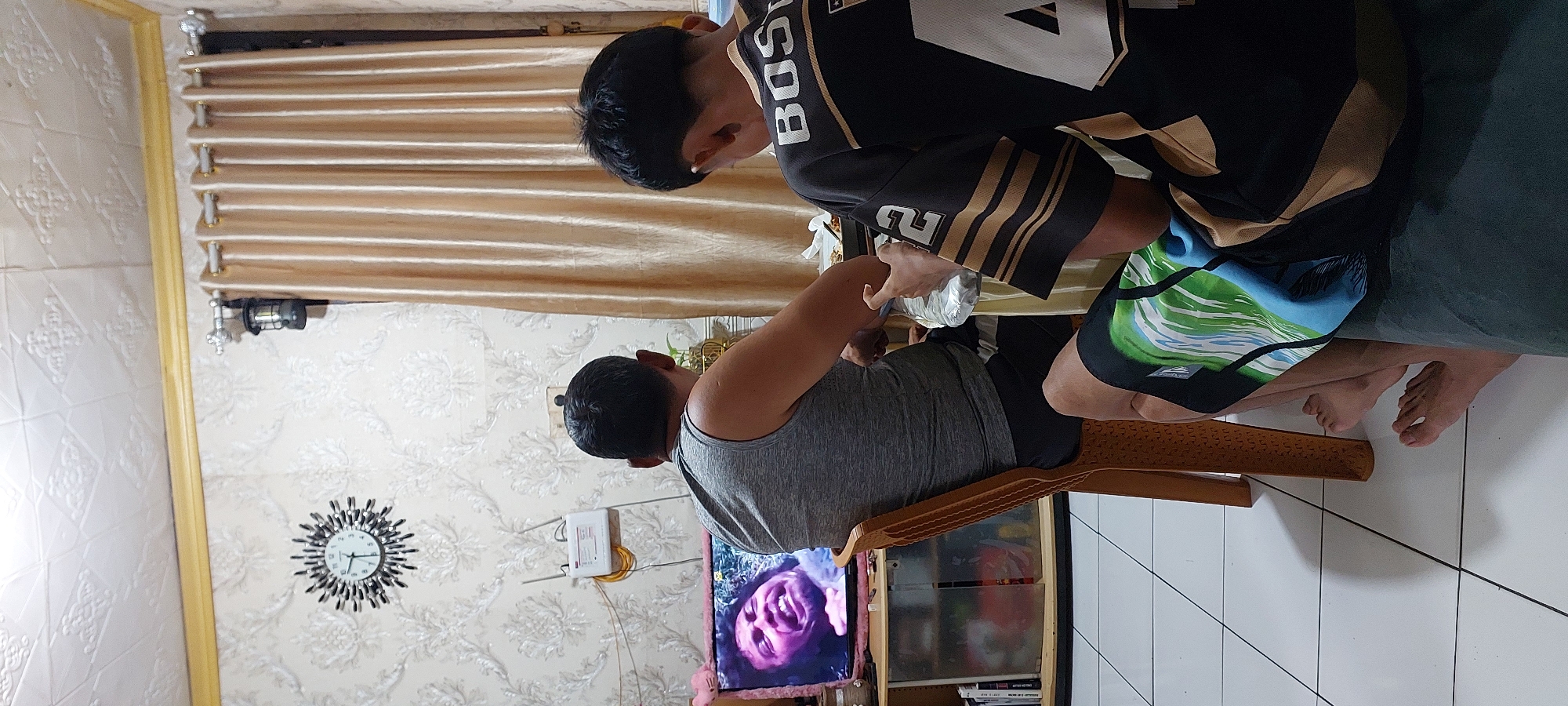 Two individuals, viewed from behind, are seated in a room, facing a television screen that displays a close-up of a person's face with an exaggerated expression. One individual is wearing a grey sleeveless shirt and black shorts, seated on a brown plastic chair. The other individual, in front, is wearing a black and gold patterned shirt with the number "4" and "2" visible, and patterned shorts. They are seated on a dark cushion. A table is positioned between them with various items on it, including what appears to be food containers. In the background, there is a wall with embossed wallpaper, a starburst clock, a wall-mounted lamp, and a beige curtain. A bookshelf with visible items is to the left of the television. The floor is tiled with white square tiles. The scene suggests a casual indoor activity, likely entertainment or a meal. The image is taken from a slightly elevated perspective. The context of Jayapura, Indonesia is not visually discernable from the image itself.