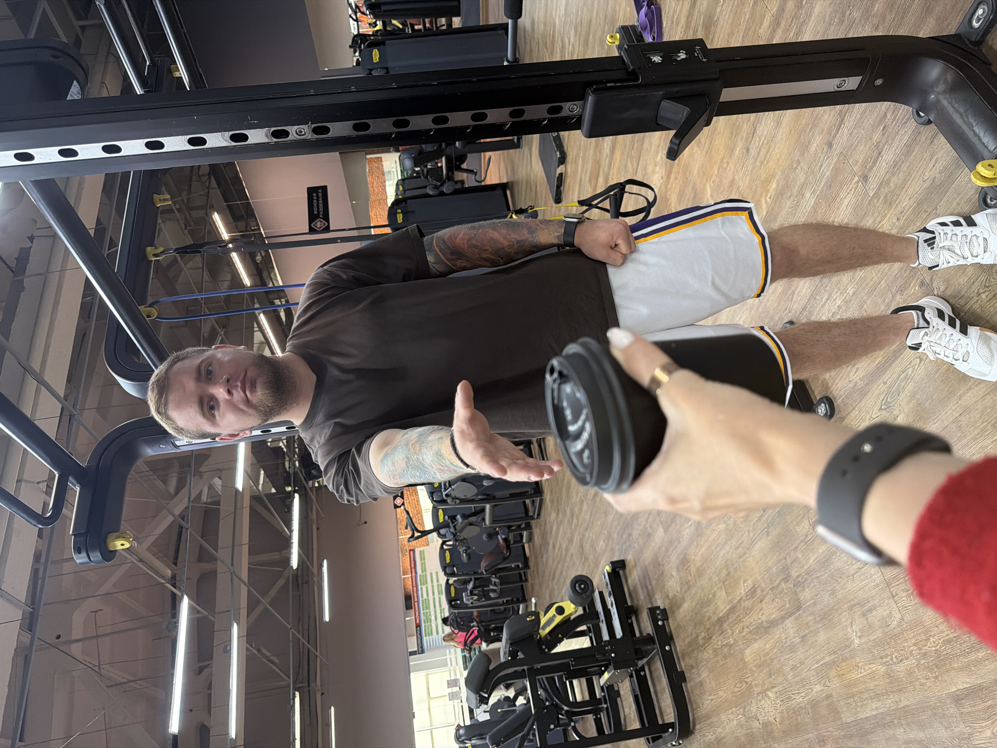 A young man with light brown hair and a beard stands in a gym, reaching out his left hand towards the camera. He is wearing a dark brown t-shirt and white shorts with yellow and purple stripes. His arms are covered in tattoos. In his right hand, he holds a black coffee cup, with the top of the cup in the foreground of the image. The gym has wooden floors and various exercise machines in the background. The lighting appears to be artificial, with fluorescent lights visible on the ceiling. The man's expression is neutral. The scene suggests an interaction where the man is either offering or receiving the coffee. The image was taken in Chaikovsky, Russia.