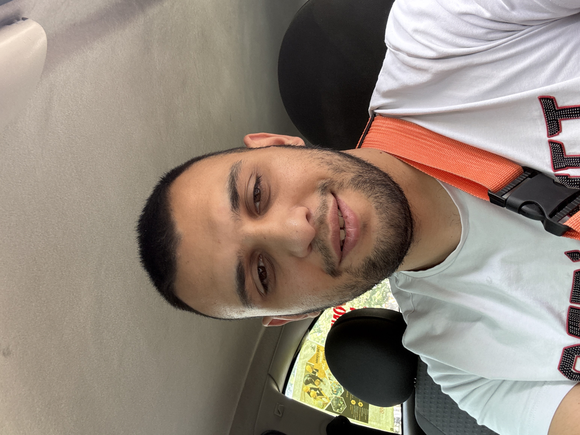A portrait of a man, taken in the passenger seat of a vehicle in Ruse, Bulgaria. He has dark hair, a short beard, and wears a white t-shirt with black and red lettering. An orange seatbelt strap is visible across his chest. The interior of the vehicle is visible, with a gray headliner and a black seat. Through the window, there is a glimpse of a paper advertising or information sheet.