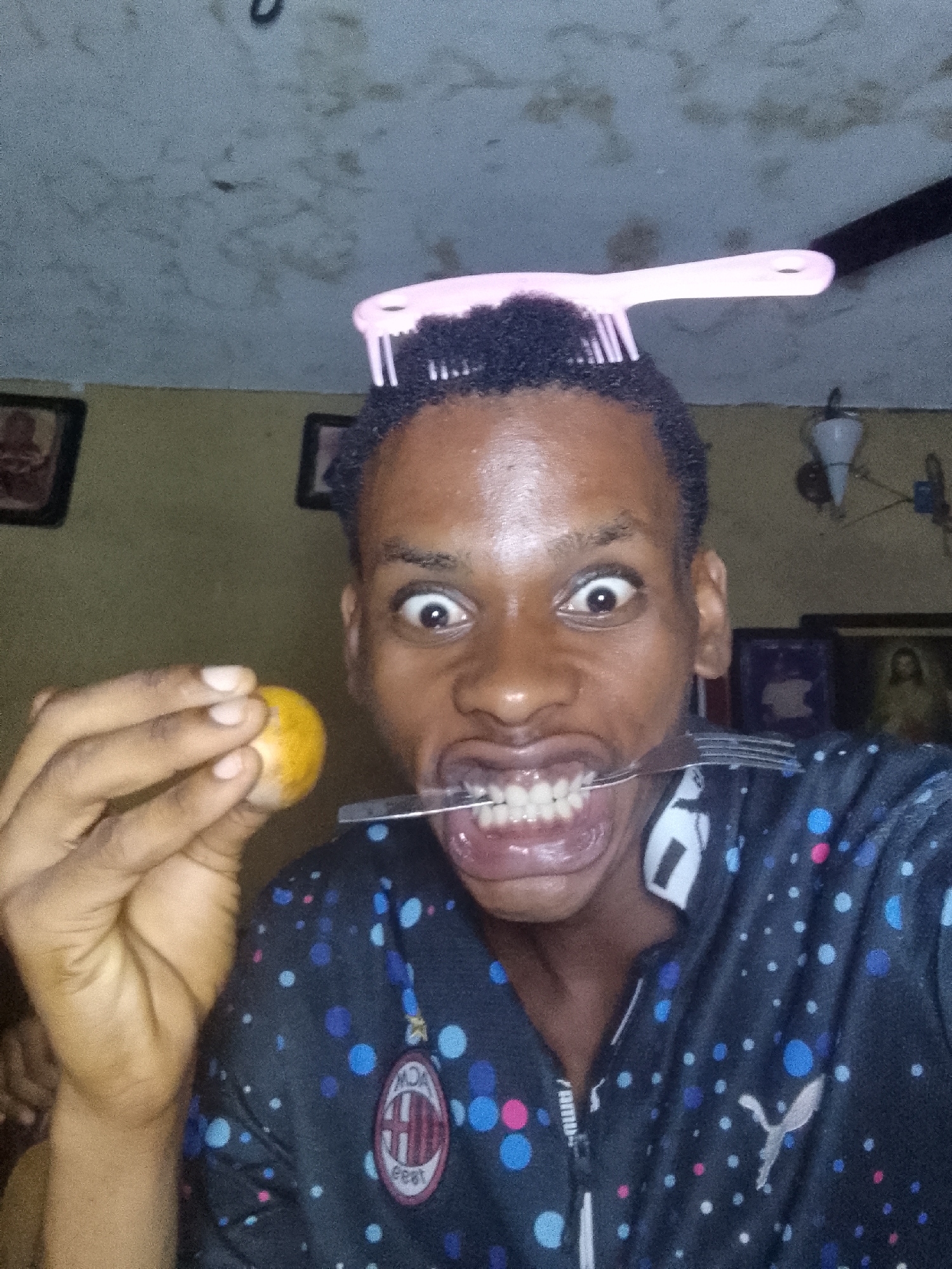 A person is posing indoors, making a face with wide eyes and baring their teeth while holding a fork in their mouth. In one hand, they are holding a small, round, yellow object. A pink comb is balanced on their head. They are wearing a dark blue jacket with a pattern of multi-colored dots and a logo on the left chest. Behind them, there is a wall with pictures hanging on it, and a ceiling with what appears to be stains or discoloration.