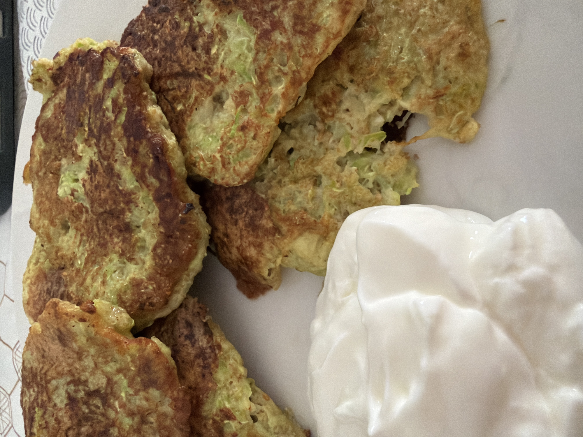 A close-up, overhead shot shows several zucchini fritters piled on a white plate, next to a dollop of white yogurt or sour cream. The fritters are golden brown and have a slightly uneven texture, indicating they are homemade. The yogurt is thick and creamy. The background is out of focus, but appears to be a patterned surface. There are no people or explicit indicators of location in Budva, Montenegro.