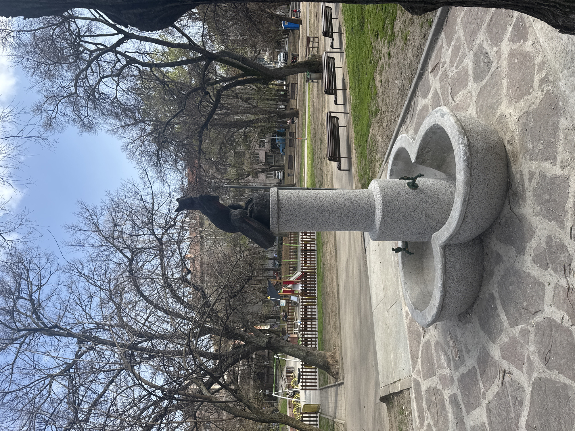 A vertical, outdoor shot reveals a park in Sofia, Bulgaria. A gray, granite water fountain stands prominently on the right with two spouts and a clover-like design. The ground around the fountain is paved with stone.
Behind the fountain, a pathway leads into the park. Park benches line the pathway, and a children's playground is visible in the distance.
Bare trees dominate the left side of the frame, their branches reaching towards a light blue sky with scattered clouds. In the background, buildings and a hint of the city can be seen.