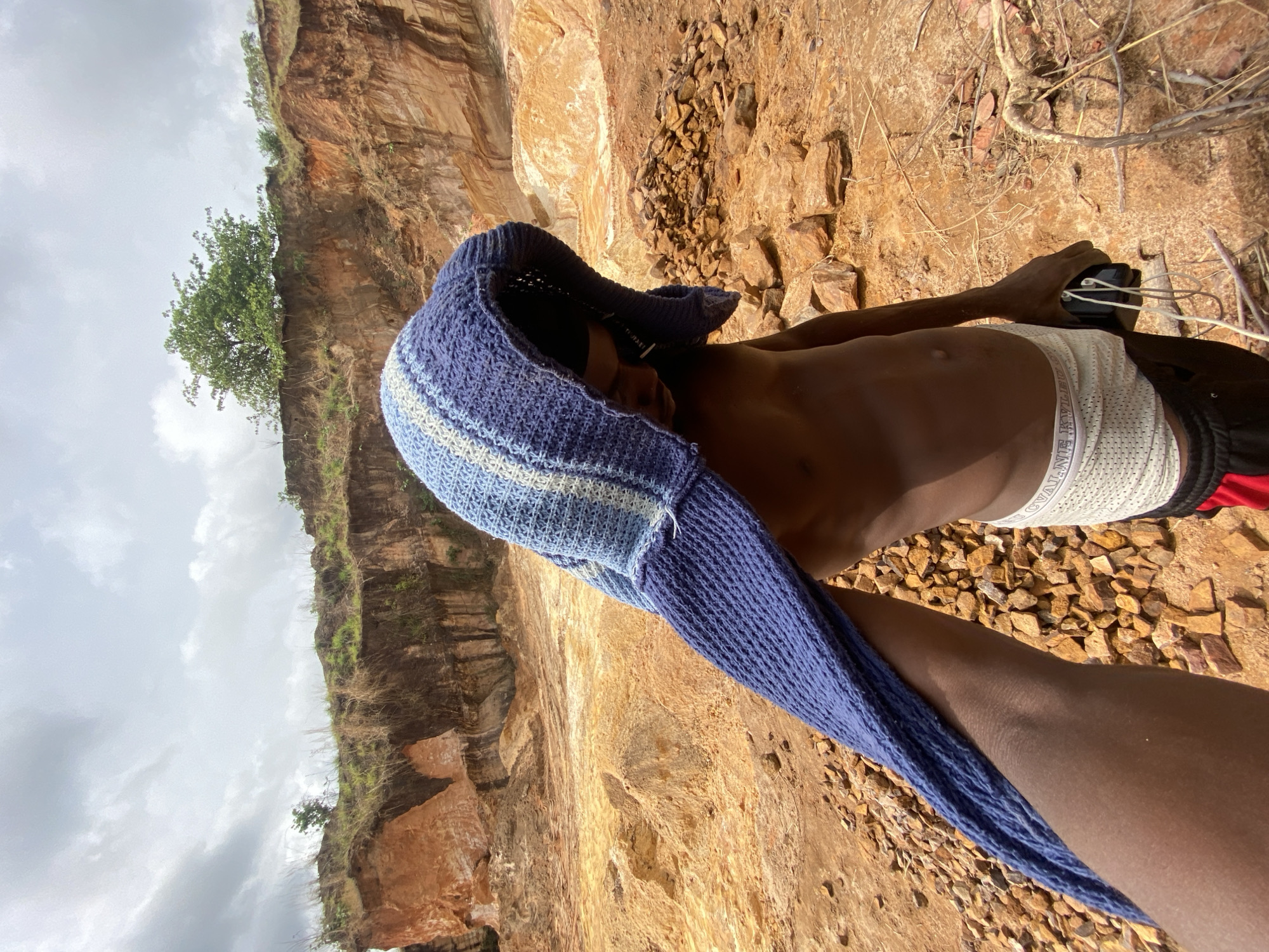 This image depicts an outdoor scene in Ufu Esioni, Nigeria, characterized by a barren, reddish-orange landscape under an overcast sky. In the immediate foreground, a dark-skinned hand from the left is visible, holding and adjusting a blue and white striped, crocheted hooded garment that covers the head of a bare-chested individual. This individual is seen from the right side, wearing white mesh underwear with a visible waistband partially reading "VAL J", over black shorts with a red stripe. The individual's right hand grasps a black electronic device with white wires dangling from it. The ground consists of reddish-orange soil and scattered small rocks. In the background, prominent, layered reddish-orange cliff faces rise, indicative of a quarry or an eroded area. Sparse green vegetation, including grasses and a single tree, grows along the top edges of these elevated landforms.