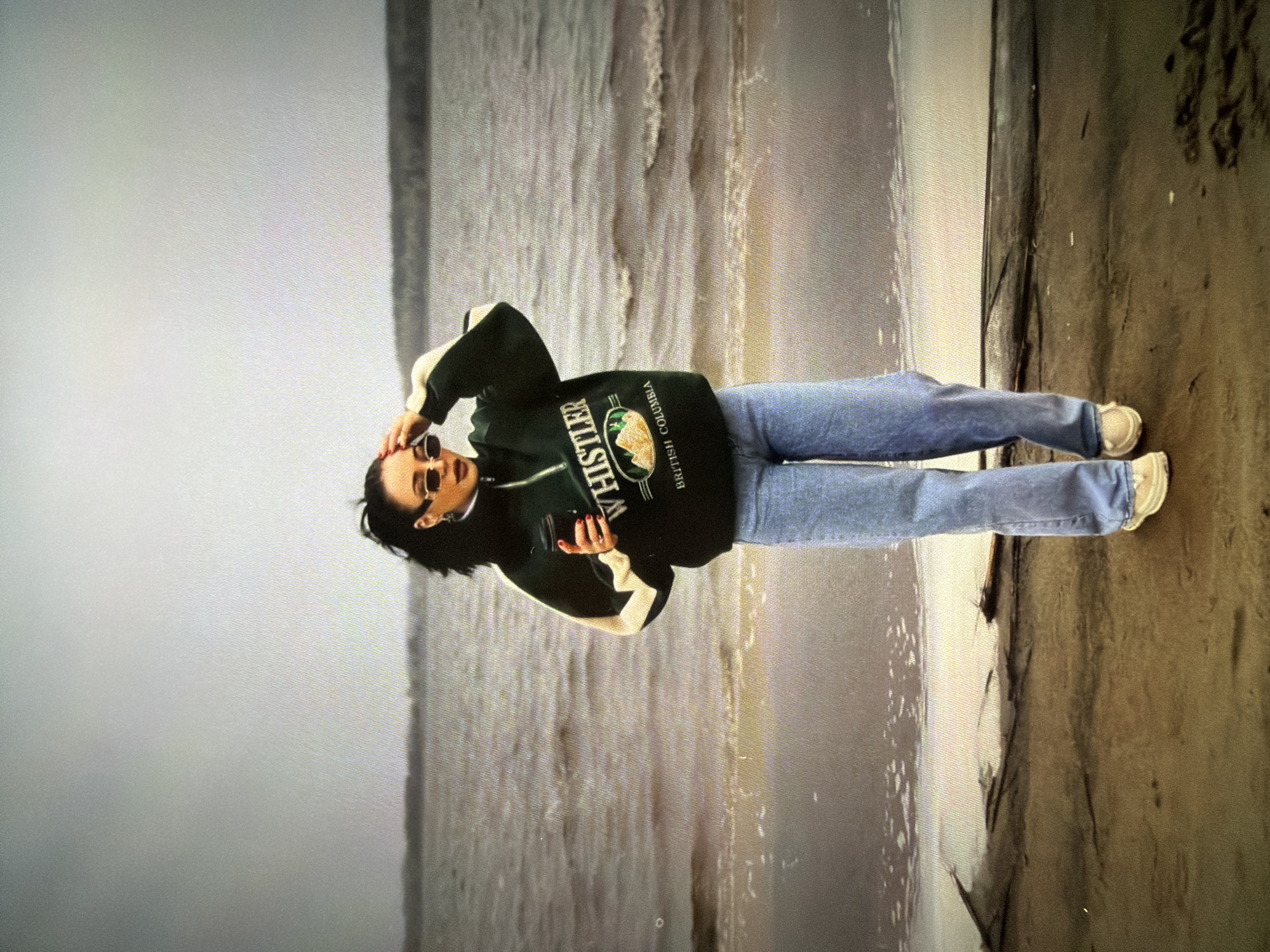 A young woman stands on a sandy shoreline with a body of water behind her. She is wearing light-wash, wide-leg jeans, a dark green half-zip sweatshirt with a "Whistler British Columbia" graphic, white sneakers, and thin, rectangular sunglasses. She holds a dark cup in her right hand and has her left hand near her face, adjusting her hair. The sky is overcast, and the water is choppy. The scene appears to be a natural landscape with no identifiable city features.