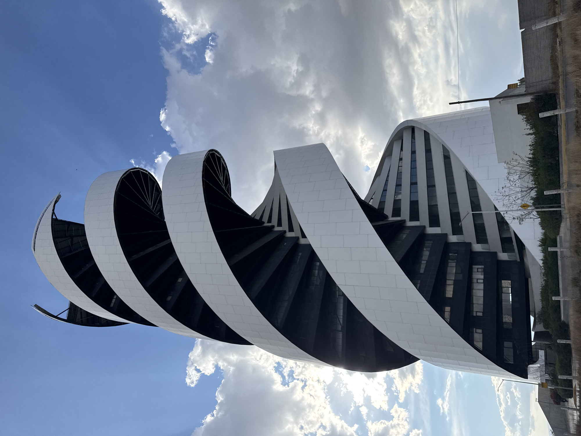 The image shows a modern architectural building located in San Bernardino Tlaxcalancingo, Mexico. The building has a distinctive design, featuring a series of spiraling or curving white bands or sections with black interior areas. These bands appear to encircle or wrap around the main structure of the building, creating a visually striking effect. The building itself is a mix of white and dark glass elements. A partly cloudy sky can be seen, with blue patches interspersed with white clouds. A typical street with bushes and other buildings can be seen in the far background.