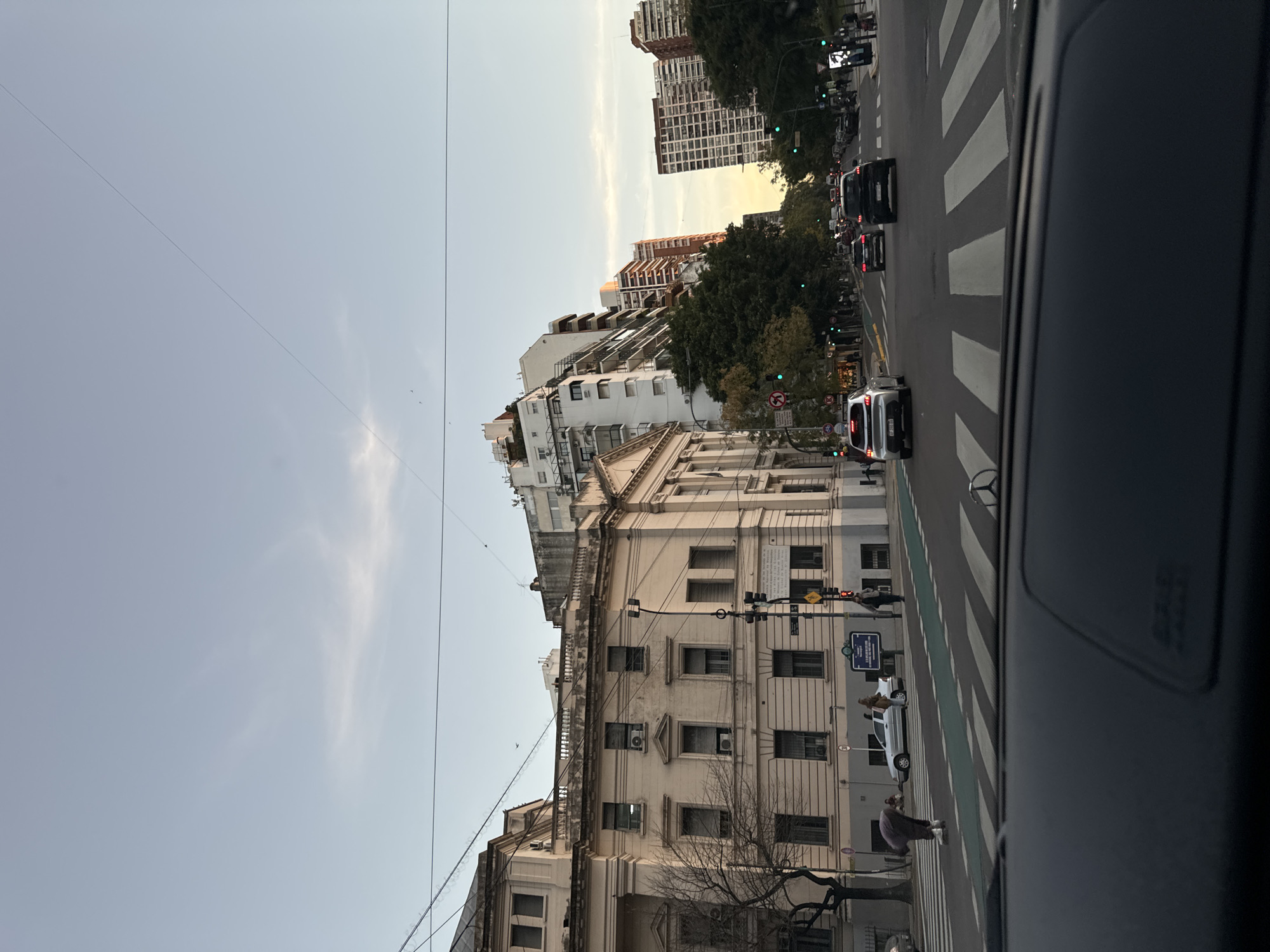 The image captures a street scene in Buenos Aires, Argentina. It's taken from inside a car, showing the dashboard and the road ahead. There's a crosswalk with pedestrians visible, and traffic signals are in operation. Buildings line the street, showcasing a mix of architectural styles. Several vehicles are present on the road. The sky is partly cloudy.