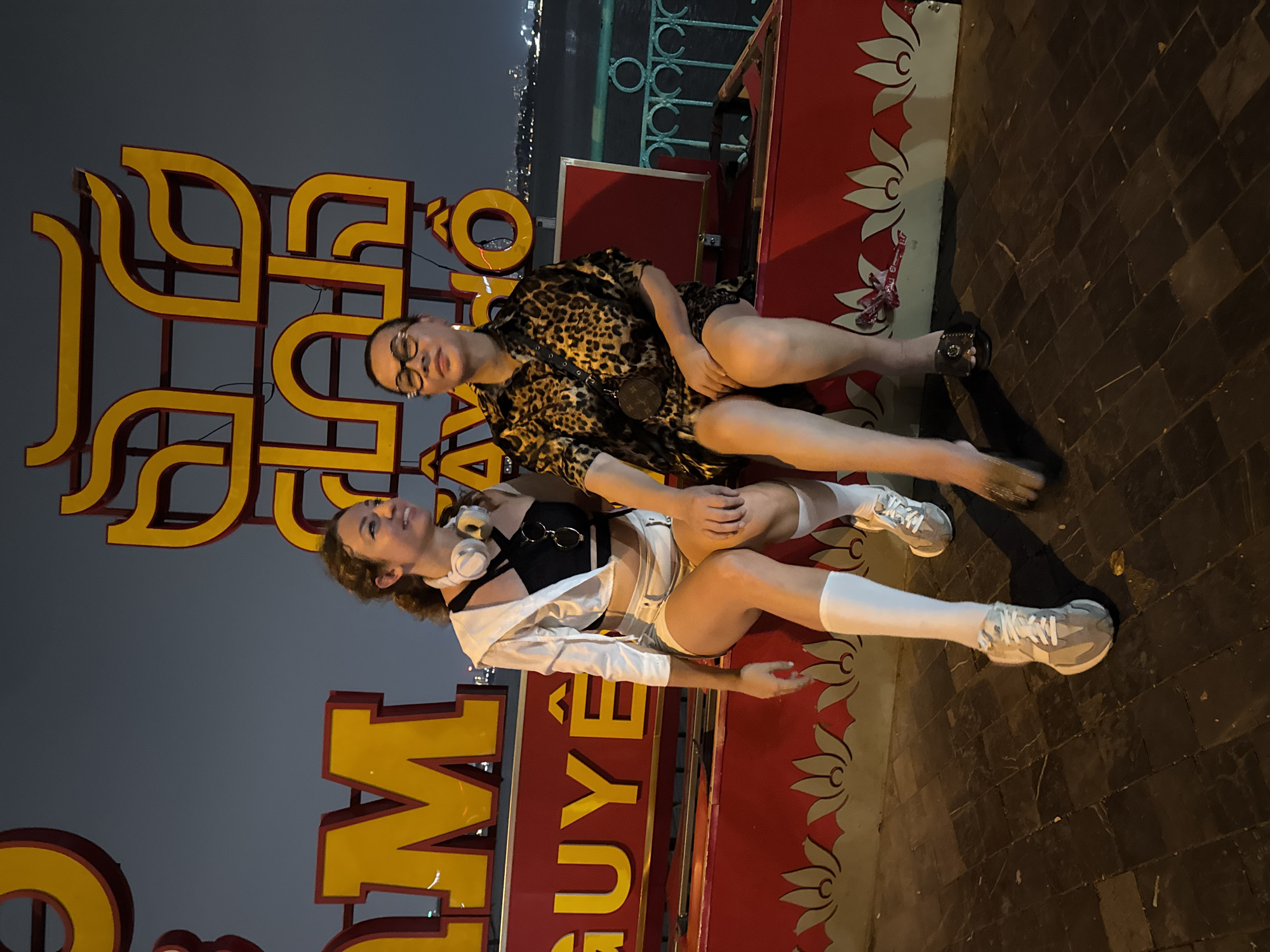 Two young women are posing for a photograph at night. The woman on the left has curly brown hair and is wearing headphones around her neck. She has on a white open shirt over a black top and white shorts. The woman on the right is wearing a leopard print dress and glasses. They are both sitting on a raised platform with a red border decorated with white floral motifs. Behind them are large yellow letters with red outlines, forming part of a sign. The ground is paved with dark square tiles. In the background, city lights are visible, suggesting an urban setting. The overall mood appears casual and playful. The visible text on the sign appears to be Vietnamese.