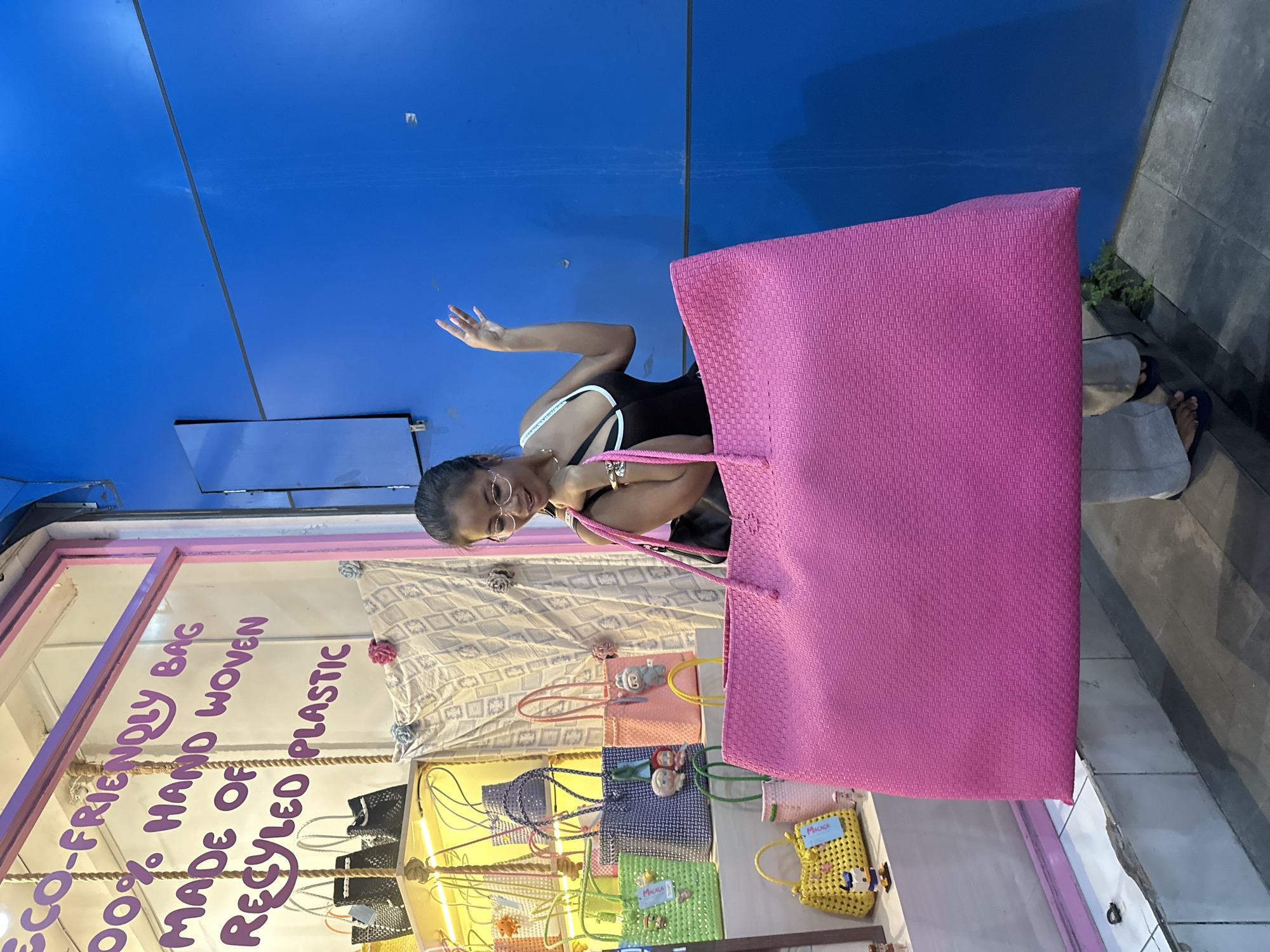 The image shows a woman in the Ubud District, Indonesia, holding a large, pink, woven bag in front of a shop window that displays various other bags. The woman has dark hair, wears glasses, a black top with white straps, and jewelry on her wrist. She is smiling and waving her hand. The shop window has a pink frame and features text promoting "eco-friendly bags," which are "100% hand-woven" and "made of recycled plastic." The bags displayed in the window come in different colors and sizes, including black, yellow, green, and blue. A blue wall is visible in the background. The ground appears to be tiled or paved.