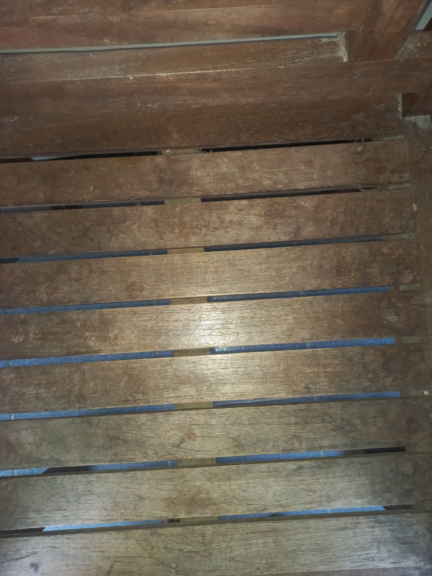 The image shows a close-up, eye-level view of weathered wooden planks, likely forming a bench or part of a seating structure, possibly in an outdoor setting. The planks are oriented horizontally and show signs of age, with visible grain, minor imperfections, and variations in color. Gaps between the planks reveal a dark, textured surface beneath, possibly a different material or shadow. The lighting suggests an ambient indoor or shaded outdoor environment, casting some subtle reflections on the wood. The scene is devoid of any people or specific identifiable landmarks within Sukoharjo, Indonesia. The focus is entirely on the material and texture of the wooden surface.