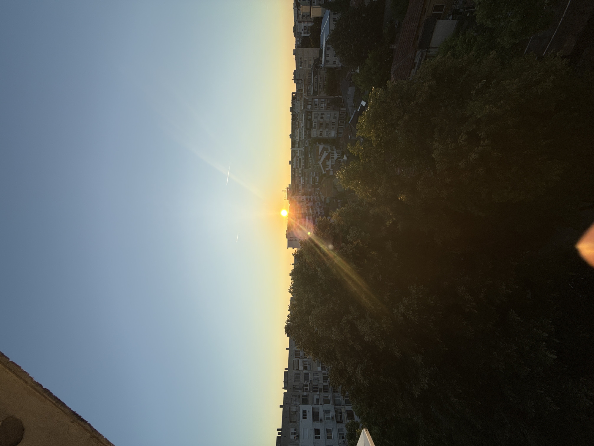 The image captures a sunrise over the city of Dobrich, Bulgaria. The sun is low on the horizon, casting a warm glow and lens flares across the scene. In the foreground, lush green trees are silhouetted against the bright sky, obscuring much of the lower city. Behind the trees, a dense urban landscape of apartment buildings and houses stretches out, with architectural styles suggesting a post-communist era. The sky is a clear gradient of blue, transitioning to yellow and orange near the sun. A faint contrail from an aircraft is visible in the upper right quadrant of the sky. The perspective suggests the photo was taken from a higher vantage point, possibly a balcony or rooftop. The overall atmosphere is serene, marked by the quiet dawn over an urban environment.