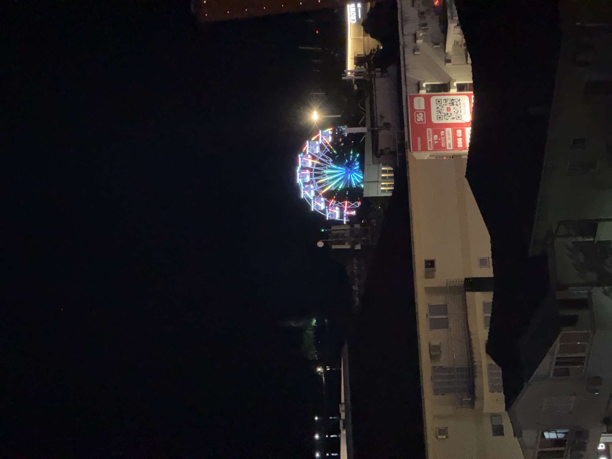 This nighttime image captures a view of Budva, Montenegro, featuring a brightly lit Ferris wheel in the background. The Ferris wheel is adorned with colorful lights, creating a vibrant spectacle against the dark sky. In the foreground, several buildings are visible, including apartments and commercial structures. One building displays a large advertisement for "5G metel" with details about a tourist SIM card offering 1 TB of data for 30 days and 500 GB. The advertisement also includes a QR code for more information. The overall scene depicts an urban environment at night, with illuminated structures and the prominent Ferris wheel suggesting a recreational area or entertainment venue.