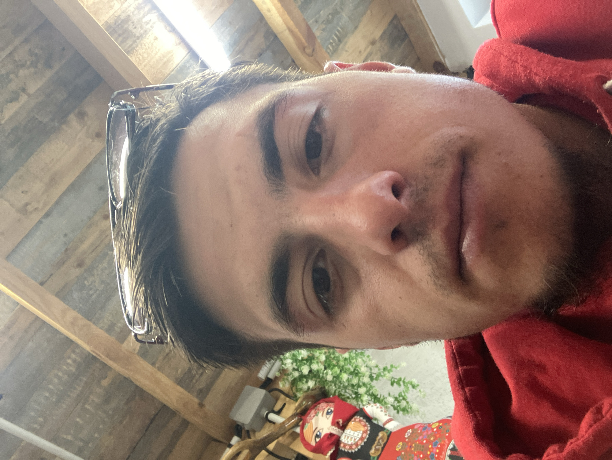 A close-up selfie of a fair-skinned male wearing a red hoodie, taken indoors. He has dark hair and stubble. Sunglasses are perched on his head. In the background, a wooden plank ceiling is visible. A doll dressed in traditional Bulgarian clothing is partially visible near a potted plant, suggesting the location might be related to Bulgarian culture, although the specific location in Komuniga, Bulgaria, cannot be determined from this image alone.