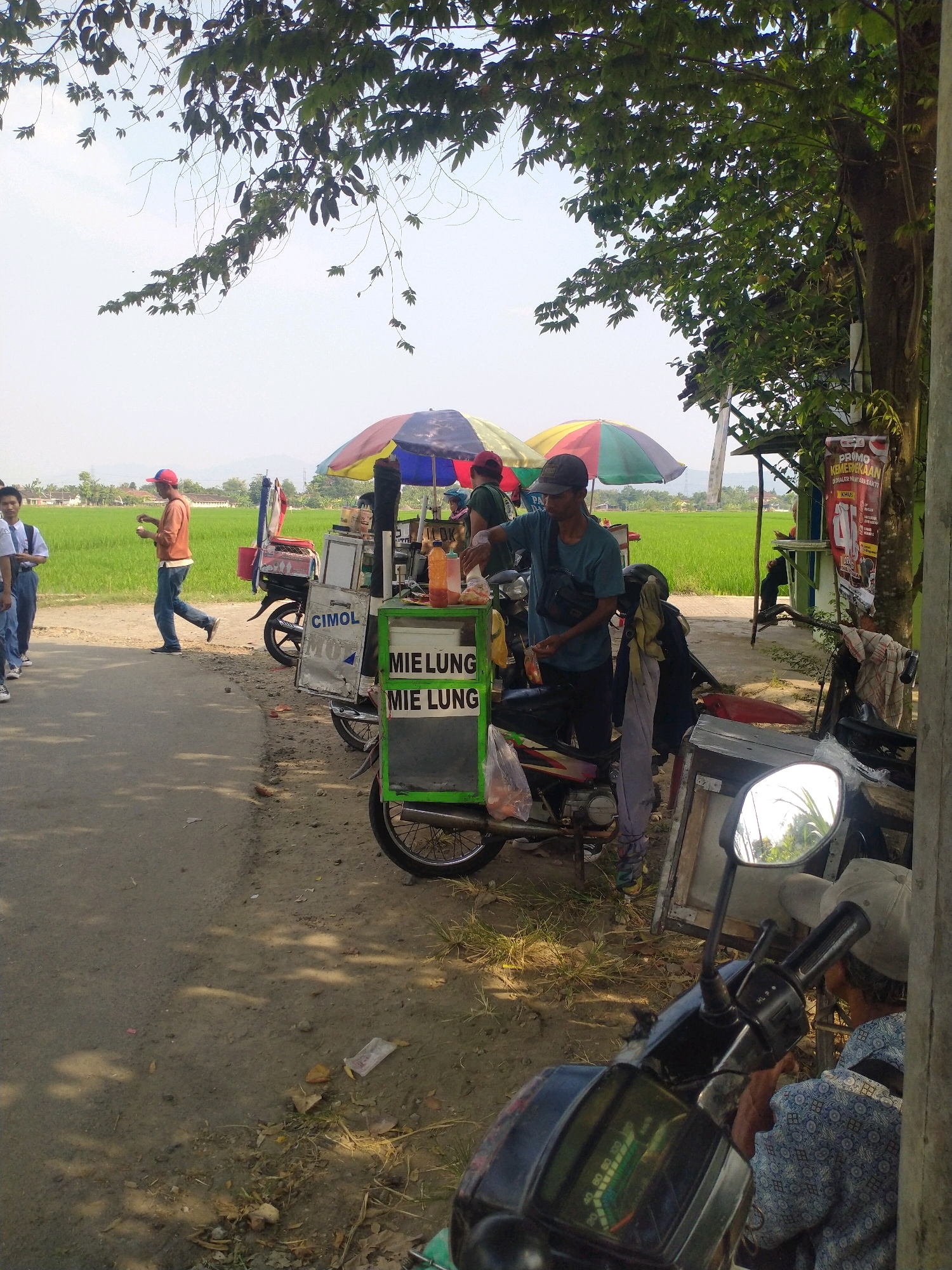 An outdoor scene depicts a roadside food stall selling "Mie Lung" and "Cimol" in Begajah, Indonesia. In the foreground, a person wearing a blue shirt and a cap is serving customers from a mobile cart. The cart is adorned with colorful umbrellas and signage advertising the food items. Behind the stall, a lush green rice field stretches towards a distant hazy horizon. Several motorcycles are parked near the stall, suggesting a bustling atmosphere. The ground is a mixture of dirt and asphalt, with some scattered debris. The overall impression is one of a vibrant local marketplace set against a backdrop of rural Indonesian landscape.