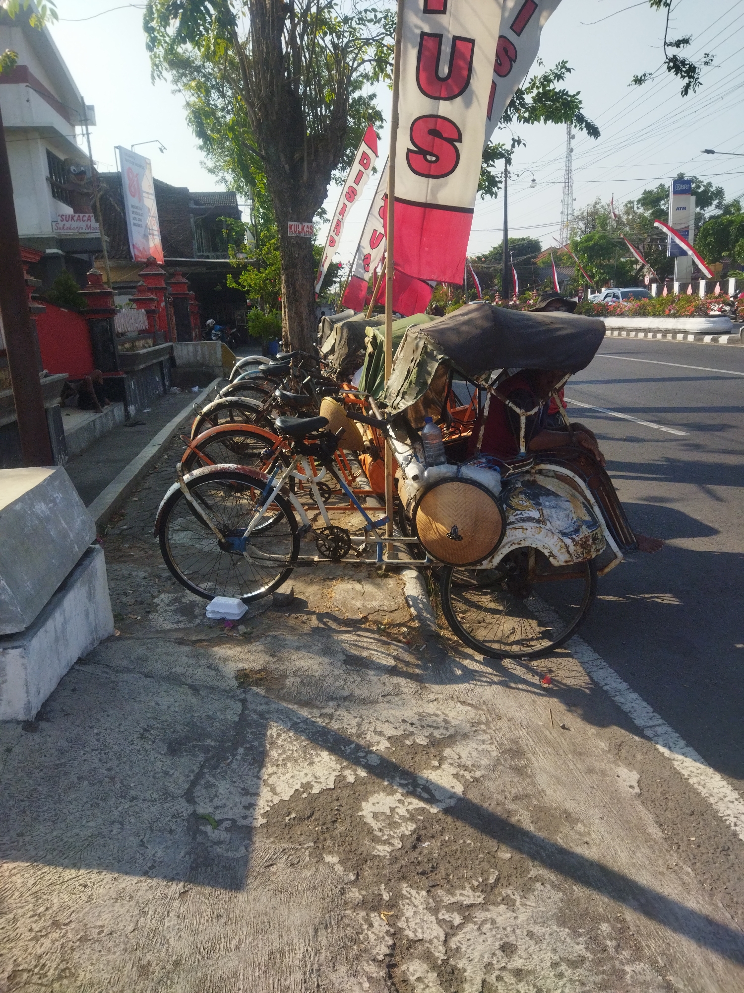The image depicts a row of parked becak, a type of three-wheeled passenger pedicab, on a sidewalk in Sukoharjo, Indonesia. The becak are lined up along a street, with a building and a tree on the left side of the frame. Several flags are visible, including red and white Indonesian flags and a banner with red lettering. One becak in the foreground is partially covered with a tarp and has a woven hat-like structure on its front wheel. The sidewalk appears to be made of concrete and is in a state of disrepair in some areas. The street is paved and has a white line marking the edge. In the background, there is a building with a sign that reads "BRI" and "ATM". The scene is bathed in sunlight, casting long shadows.