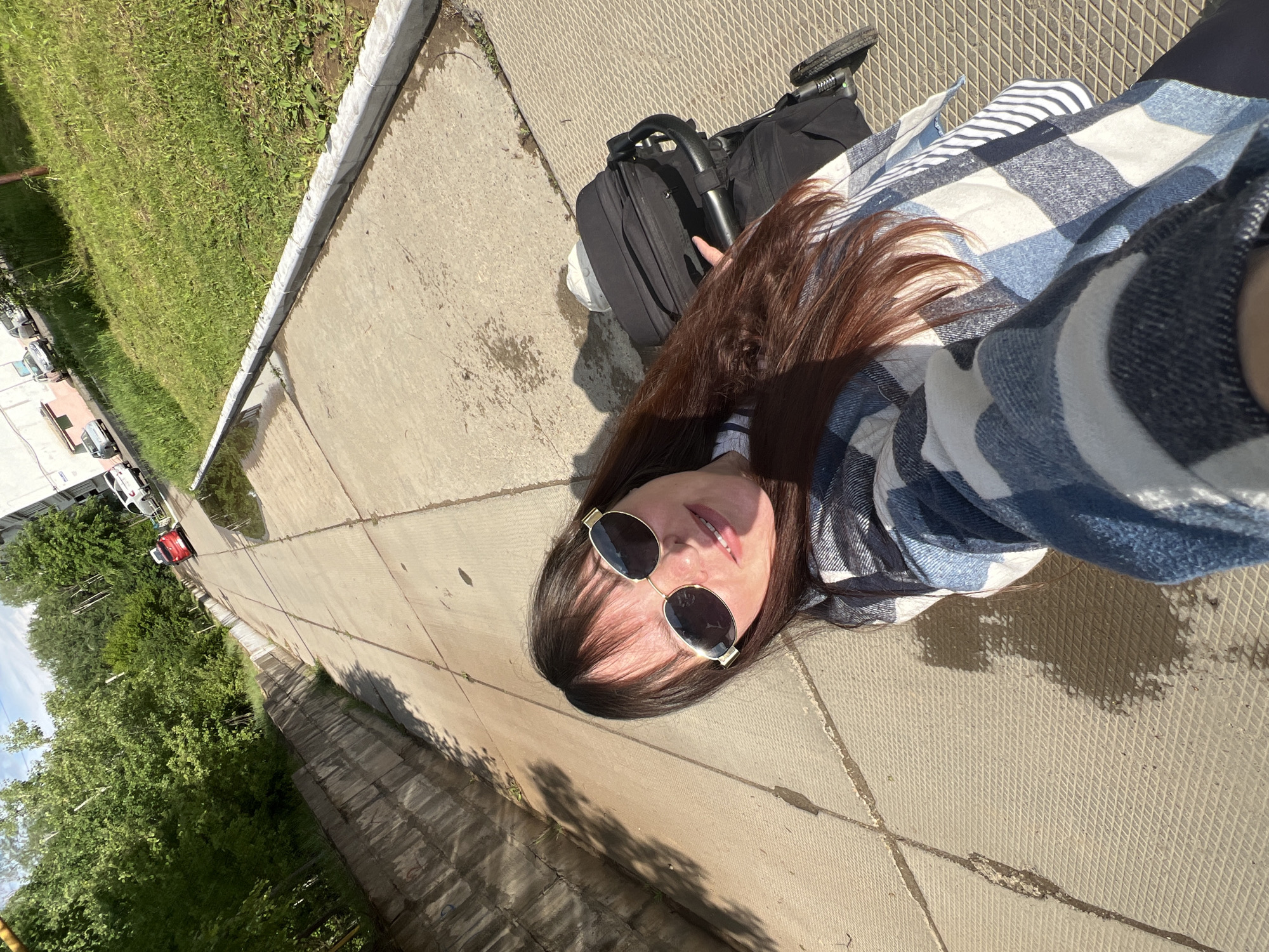 A selfie-style photograph shows a woman lying on a concrete sidewalk next to a grassy area and road. A stroller is next to her. She is wearing sunglasses and a blue and white plaid shirt. The photo appears to be taken in Noviy, Russia, based on the location information provided.