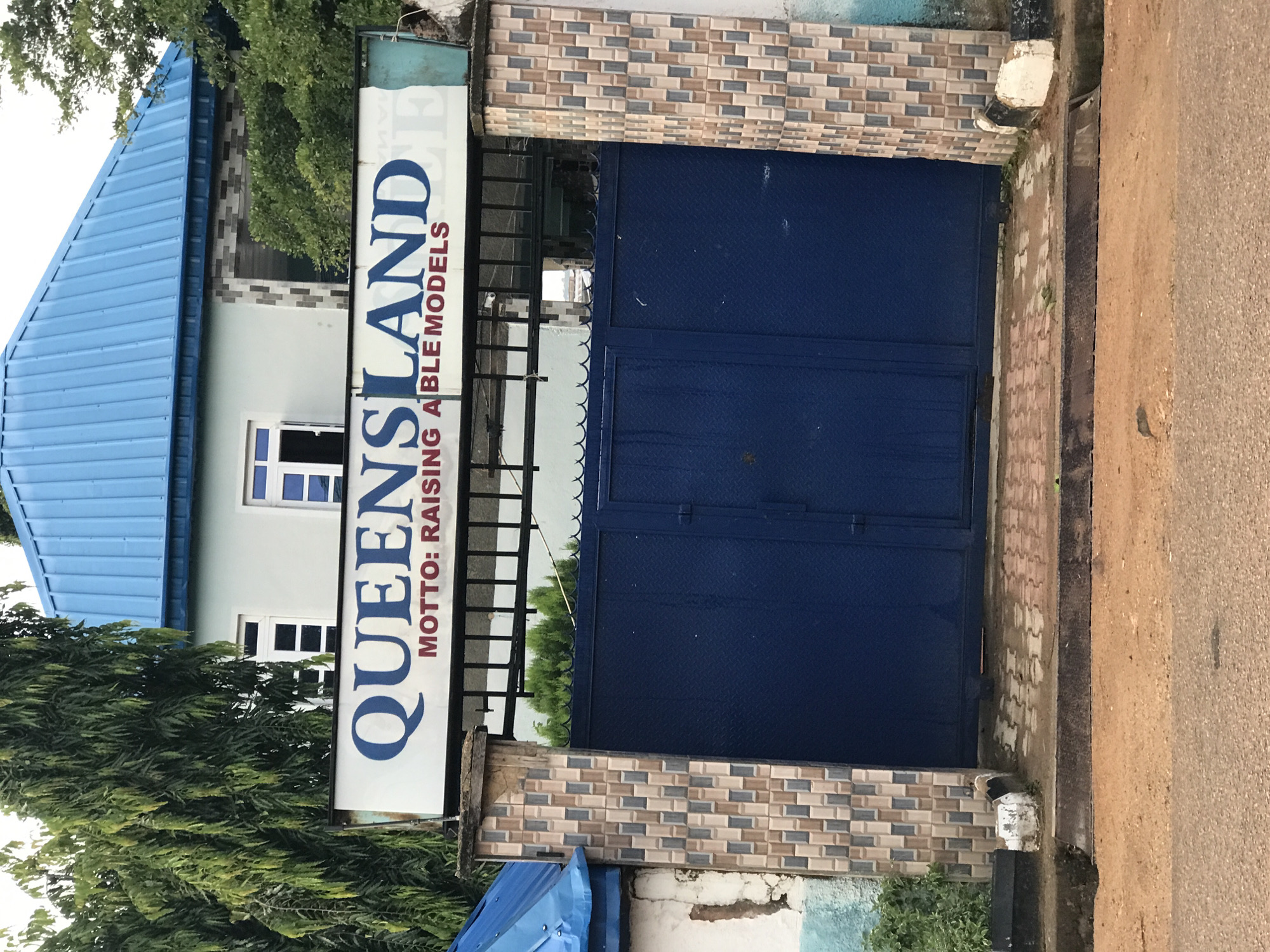 The image shows the entrance to Queensland, located in New Stadium, Nigeria. A large blue metal gate is the prominent feature, flanked by decorative tiled pillars on either side. Above the gate, a white signboard displays "QUEENSLAND" in large blue lettering, with the motto "MOTTO: RAISING ABLE MODELS" in smaller red text below. Behind the gate and sign, a light blue building with a corrugated blue roof is visible. Tall, leafy trees are positioned to the left of the building, and lush green foliage can be seen to the right. The ground in front of the gate is paved with interlocking concrete blocks. The overall scene suggests a formal establishment or institution.