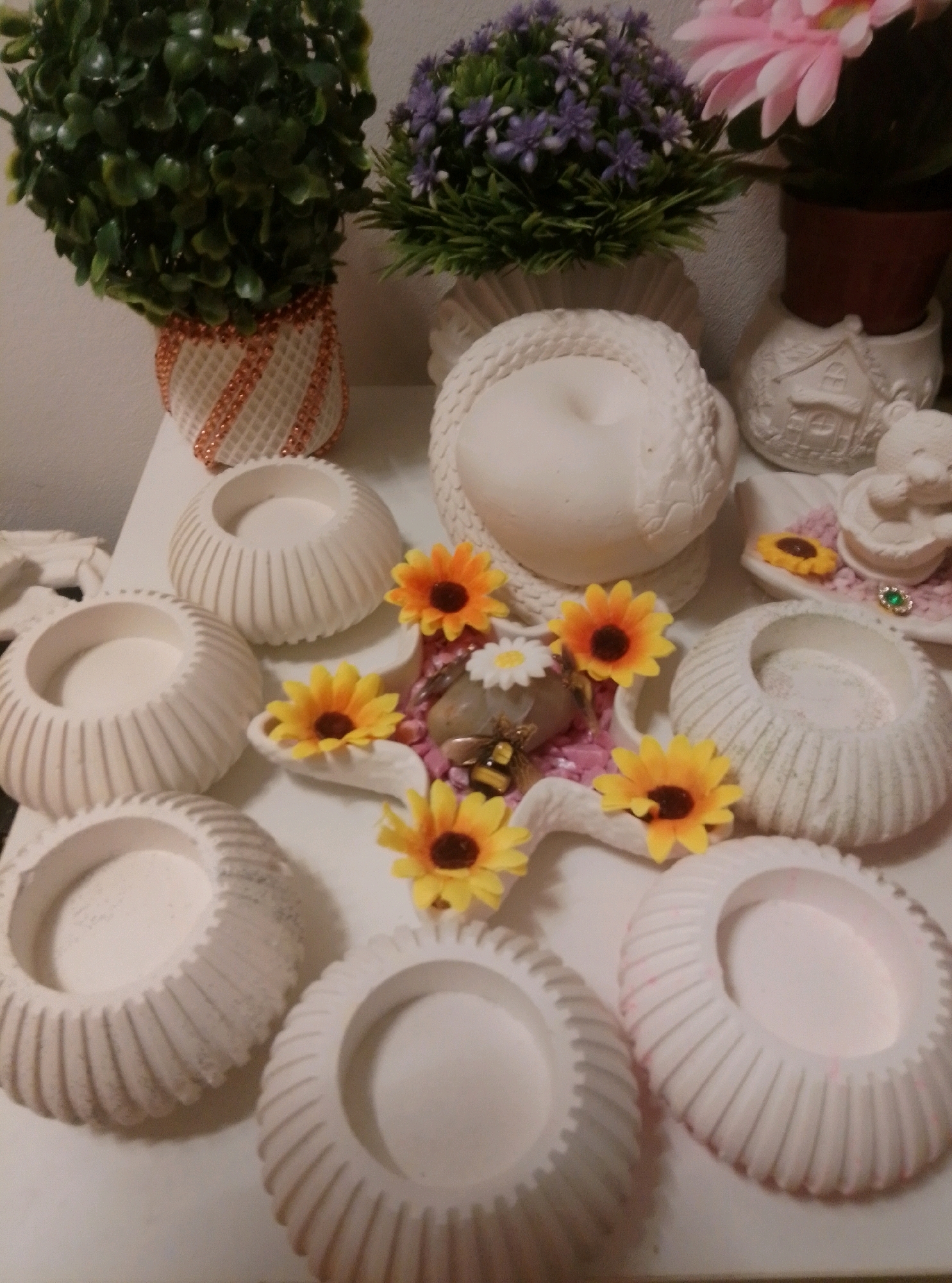 This image is a close-up shot of a collection of decorative items, possibly for crafting or display, set against a light-colored surface. The items include several white, ribbed ceramic or plaster containers, some shaped like shallow bowls or holders. Among these are artificial sunflowers with orange centers and white daisies. A decorative bee figurine is also visible, positioned near some of the flowers. In the background, there are two artificial plants: one with lush green foliage in a textured pot adorned with copper-colored trim, and another with purple flowers and green leaves. To the right, a pink artificial flower sits in a brown pot. A white ceramic house-shaped object and a small white figurine resembling a cherub or animal are also present. The overall impression is of a still life arrangement, with a focus on decorative elements and artificial flora. The image does not contain any identifiable landmarks or features that would allow for localization to Wedel, Germany.