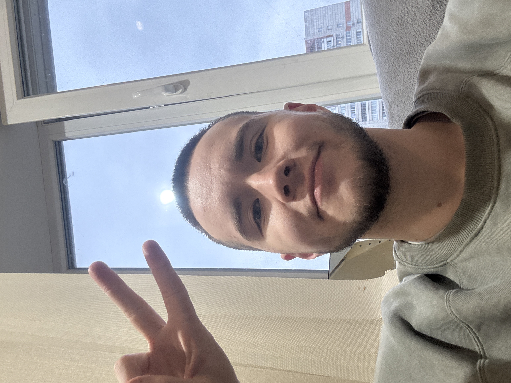 A medium-shot, selfie photograph shows a young, light-skinned man holding up a "peace sign" with his left hand. He has short, dark hair and a beard, and he is wearing a light grey sweater. Behind him is an open window with a view of a building in Kazan, Russia, suggesting the image was taken indoors during daytime. The sky is visible through the window.