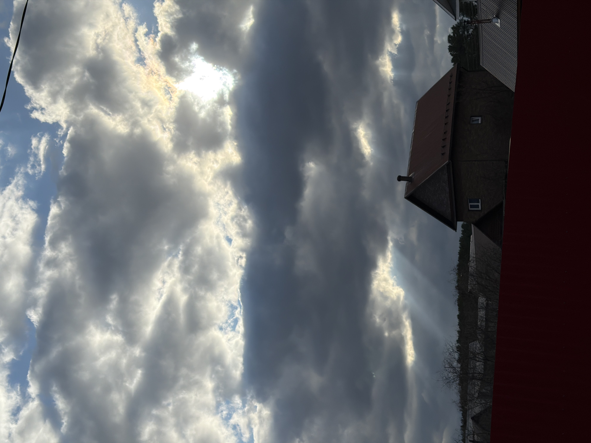 The image captures a dramatic sky dominating the upper two-thirds of the frame, showcasing a mix of thick, dark grey clouds and brightly illuminated white and golden clouds. The sun breaks through in several areas, creating brilliant patches of light and visible crepuscular rays that pierce through the cloud layers. Patches of blue sky are visible in the upper left and center left, contrasting with the heavier cloud cover. A thin, dark diagonal line, possibly a power line, traverses the upper left corner.
In the lower right portion of the image, a building with a dark facade, possibly brick or siding, and a reddish-brown gabled roof is prominent. It features a chimney on one end and two visible rectangular windows. Behind this building, a dense line of bare trees, characteristic of a forest or wooded area in late autumn or winter, stretches across the horizon. Further back, another darker structure is partially visible.
The scene depicts a natural landscape with some architectural elements, likely in a rural or semi-rural setting in Chaikovsky, Russia. The dramatic lighting suggests either late afternoon or early morning, with the low sun creating strong contrasts between light and shadow. There are no people visible, and no specific activity or event is taking place beyond the natural progression of light and clouds. No visible text is present.
