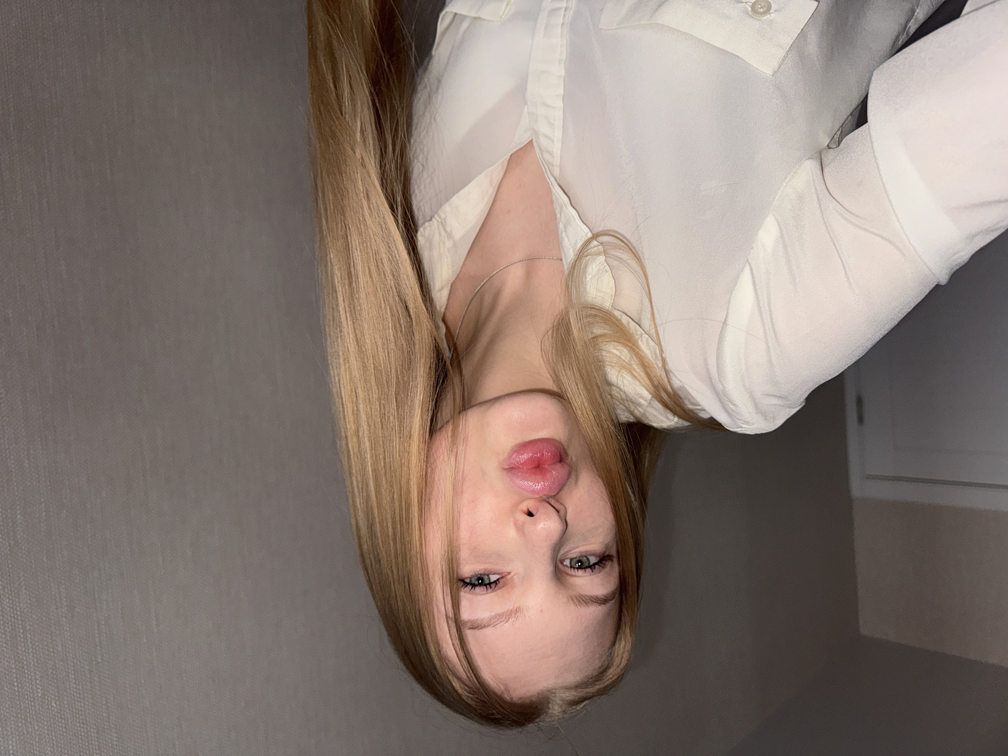 A young woman with long blonde hair and blue eyes is posing for a selfie. She is wearing a white button-down shirt and a delicate silver necklace. Her lips are puckered in a playful expression. The background consists of a gray wall. The location is not identifiable as Perm, Russia based on the visual information provided.