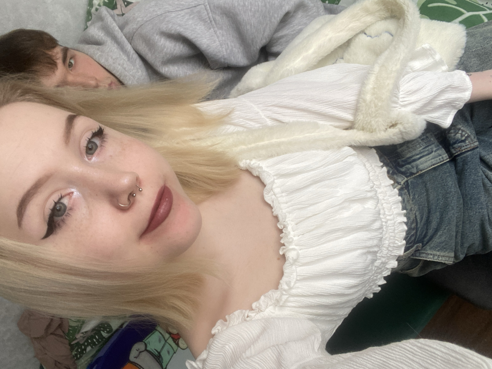 A young woman with blonde hair is lying down next to a person wearing a grey hoodie. The woman is wearing a white ruffled top and jeans, and has a nose piercing. A white fluffy bag is draped over her. The background is a bed or couch with patterned bedding. The location is not discernible from the image.