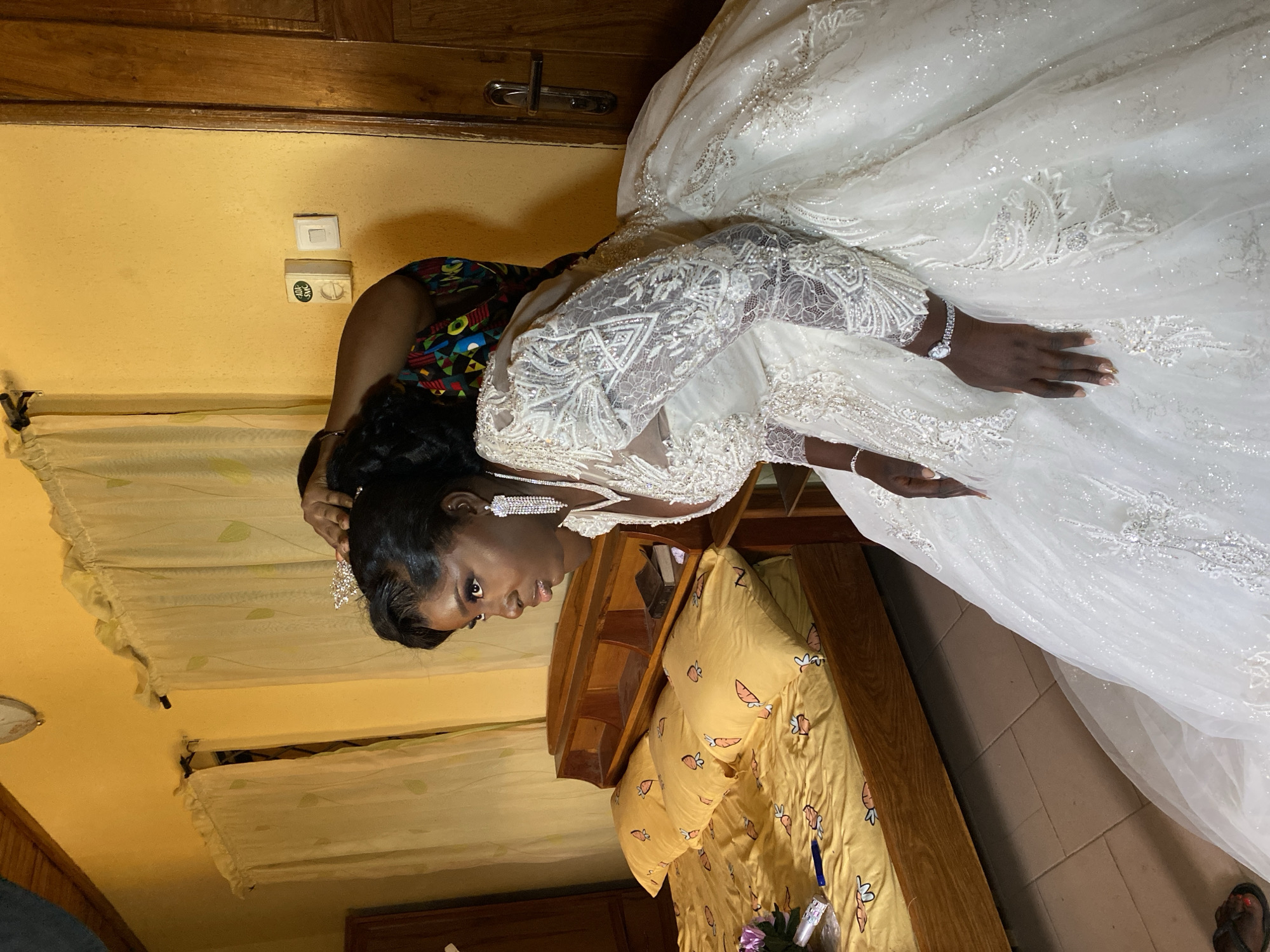 A woman is adjusting a bride's hair in a bedroom setting in Vom, Nigeria. The bride is wearing a white, embellished wedding gown, tiara, and long earrings. The other woman is wearing a brightly patterned top. The bedroom has yellow walls, a bed with yellow bedding with cartoon-like prints, and light curtains. The scene appears to be a pre-wedding preparation moment.