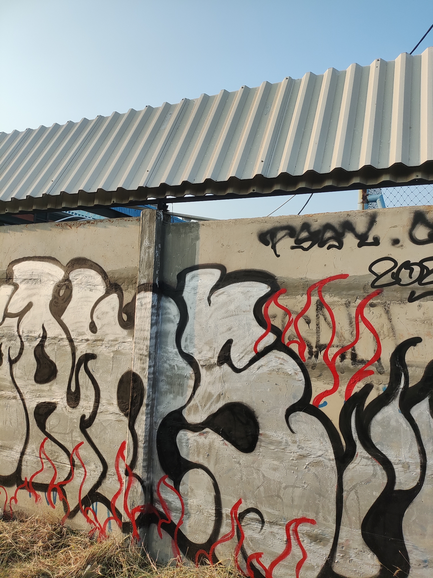 In East Jakarta, Indonesia, a section of a concrete wall is covered in graffiti. The graffiti features large, stylized black and white figures, with red flame-like accents. Above the wall, a corrugated metal roof extends diagonally across the frame, supported by unseen structures. The sky is clear and blue. Dry grass and weeds are visible at the base of the wall.