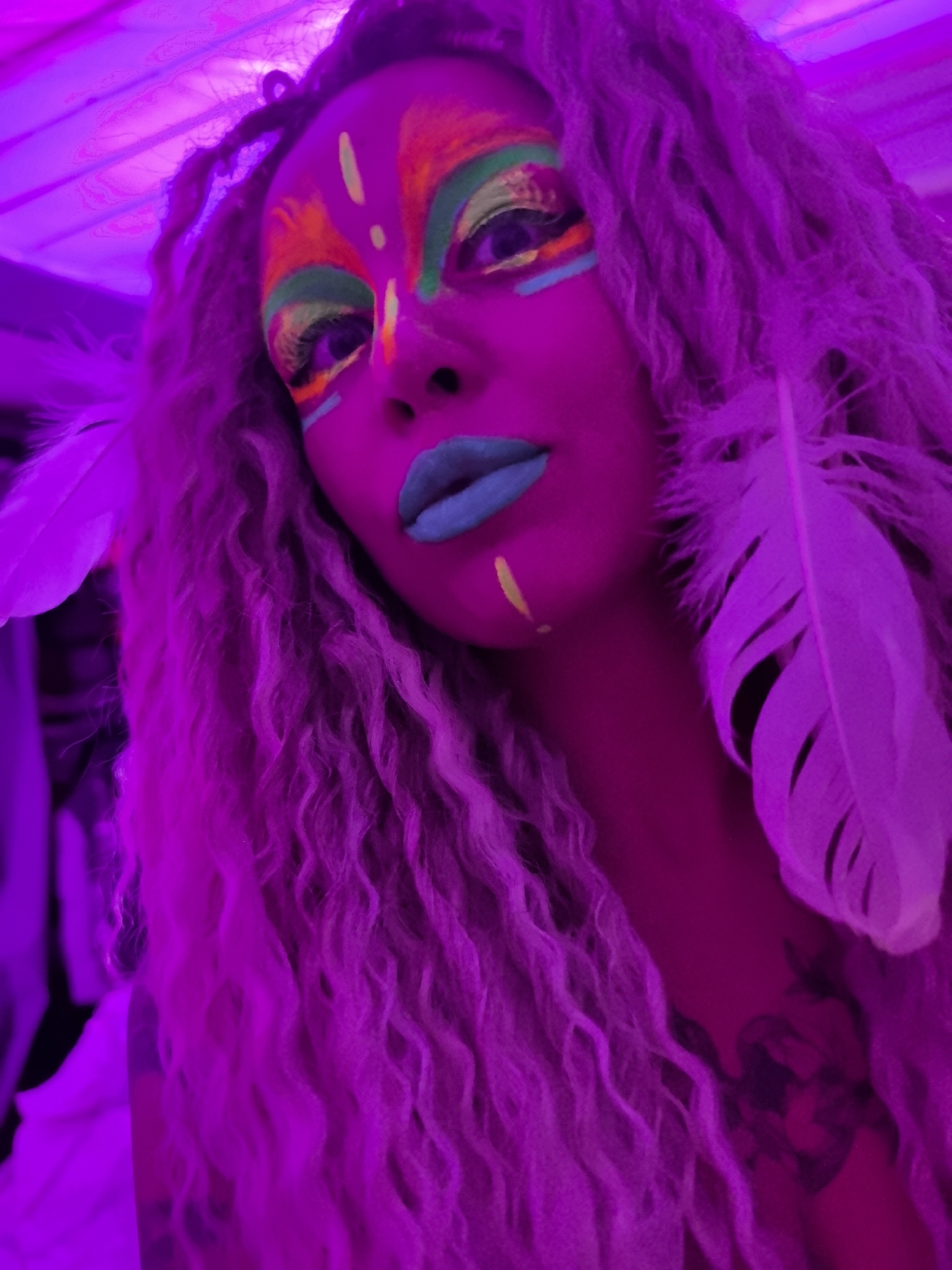A low-angle close-up shot shows a person with long, curly hair illuminated by purple light. The person's face is adorned with colorful makeup, including neon green and orange designs on the forehead and around the eyes, as well as blue lipstick. A single yellow stripe is painted on the cheek. Two white feathers are positioned near the person's face, one on each side. A tattoo is visible on the chest. The location is not identifiable as Noviy, Russia based on the provided image.