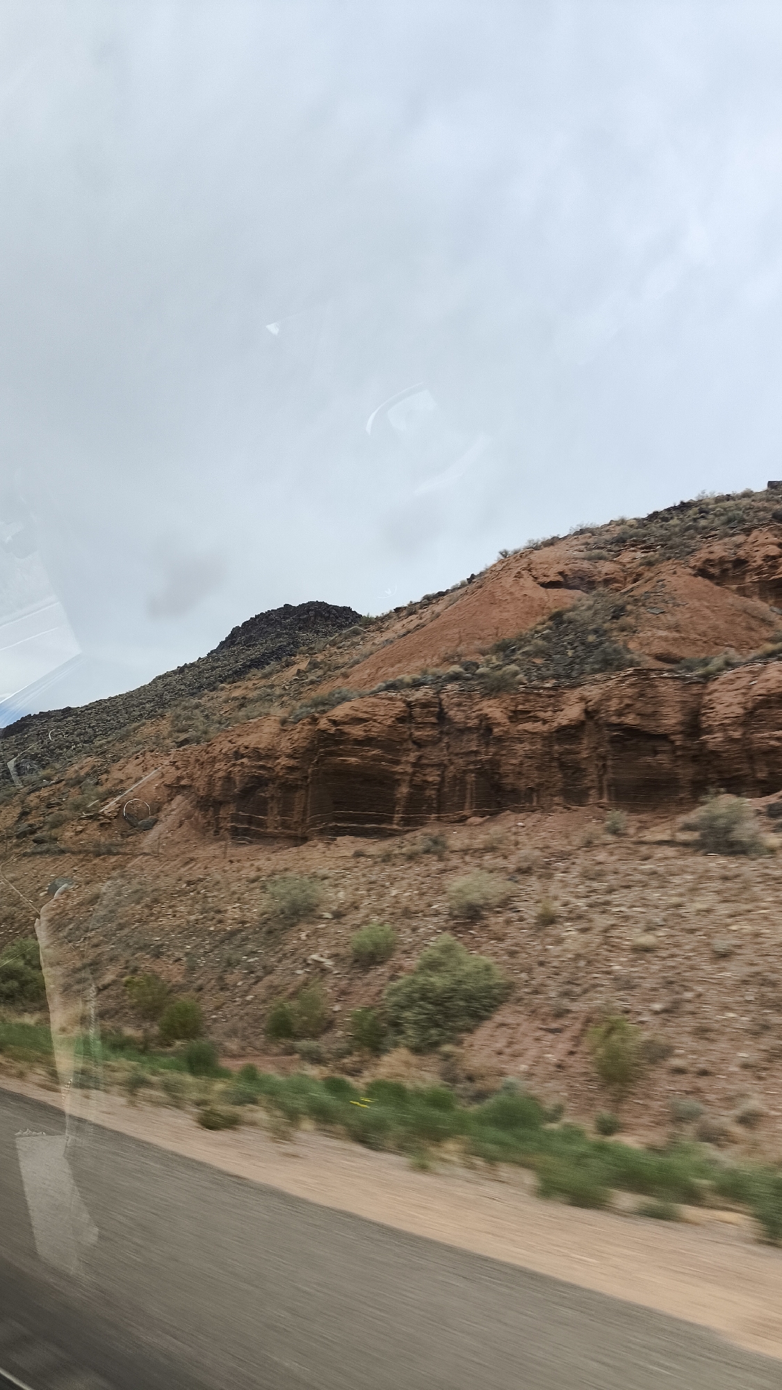 The image shows a side view of a mountainous landscape with red rock formations and sparse vegetation. The sky is overcast with a light gray hue. In the foreground, a blurred asphalt road is visible, suggesting the photograph was taken from a moving vehicle. The overall scene depicts a desert environment with rugged terrain.