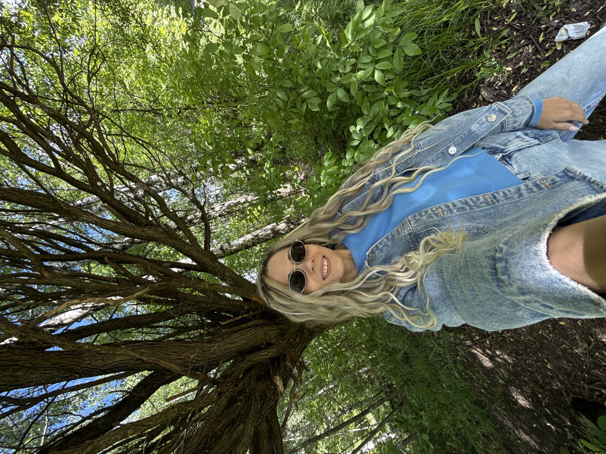 A young woman with long, blonde, wavy hair is the primary subject of a photograph. She is lying on her side in an outdoor setting, possibly a park or wooded area. She wears round sunglasses, a blue sweater, and a denim jacket. Her left hand is visible near her torso.
The woman is situated near the base of a tree with exposed, gnarled roots and branches that extend upward and to the left. Sunlight filters through the foliage, creating a dappled effect. Green foliage and vegetation surround her, with some growing in close proximity to her body. The ground appears to be covered in dirt, leaves, and small twigs.
Based on the specified location, the scene could be in a natural or landscaped area within Шебунино, Russia, where trees and greenery are prevalent.