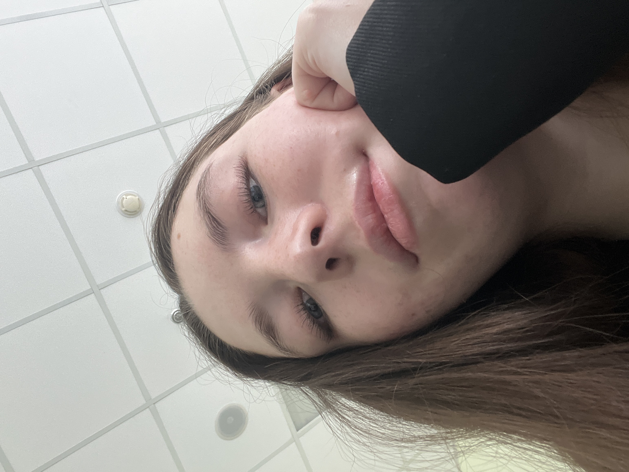 A close-up, low-angle shot focuses on a fair-skinned individual with light blue eyes and brown hair. The person's face is angled towards the camera, with their hand, wearing a black-sleeved garment, touching the side of their face near the ear. The background consists of a white ceiling with a grid pattern, punctuated by circular light fixtures. The location is not discernible as Moscow, Russia.