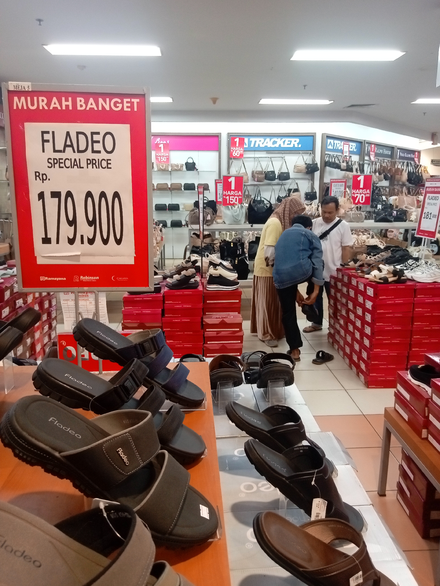 The image shows the interior of a shoe store, likely in Batam, Indonesia, based on the prominent display of the brand "Fladeo" and the pricing in Rupiah. In the foreground, several pairs of Fladeo brand sandals are displayed on a wooden shelf, with some in black and others in gray and brown. Behind these, a large red and white sign advertises "MURAH BANGET" (very cheap) and "FLADEO SPECIAL PRICE" with a price of Rp. 179,900. Further back, a man is helping a woman try on a sandal while another woman looks on. The background is filled with shelves displaying various brands of shoes and handbags. There are also stacks of shoe boxes, predominantly red, throughout the store. The overall impression is a busy retail environment with a focus on discounted footwear.