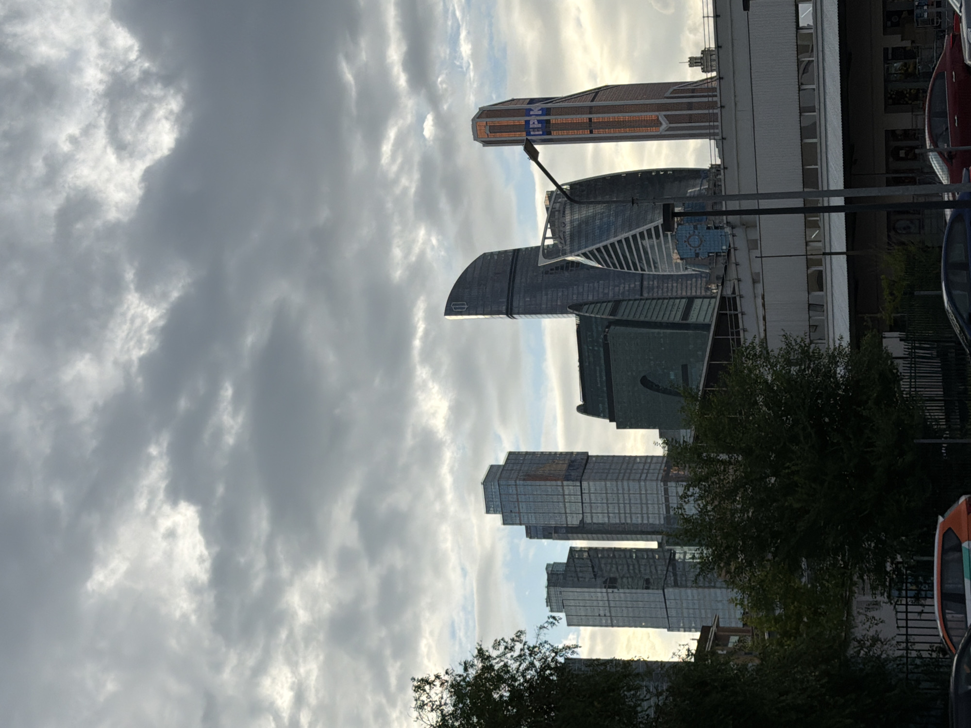 The image shows a view of the Moscow City business district with a cloudy sky. In the foreground, there is a white building with a row of windows and a metal roof. A dark-colored lamppost stands in front of it. To the left of this building, there are dark green trees and foliage, with a glimpse of a dark fence and a partial view of cars.
Dominating the background are several modern skyscrapers, characterized by their glass facades and geometric shapes. The tallest buildings include two rectangular, glass-covered towers on the left, and a cluster of more uniquely shaped towers to the right, including a curved dark grey one and a distinctively striped copper-colored tower with "IPK" signage. The sky is filled with a mix of grey and white clouds, with some patches of brighter light suggesting the sun trying to break through. The overall impression is an urban landscape under a dramatic sky.