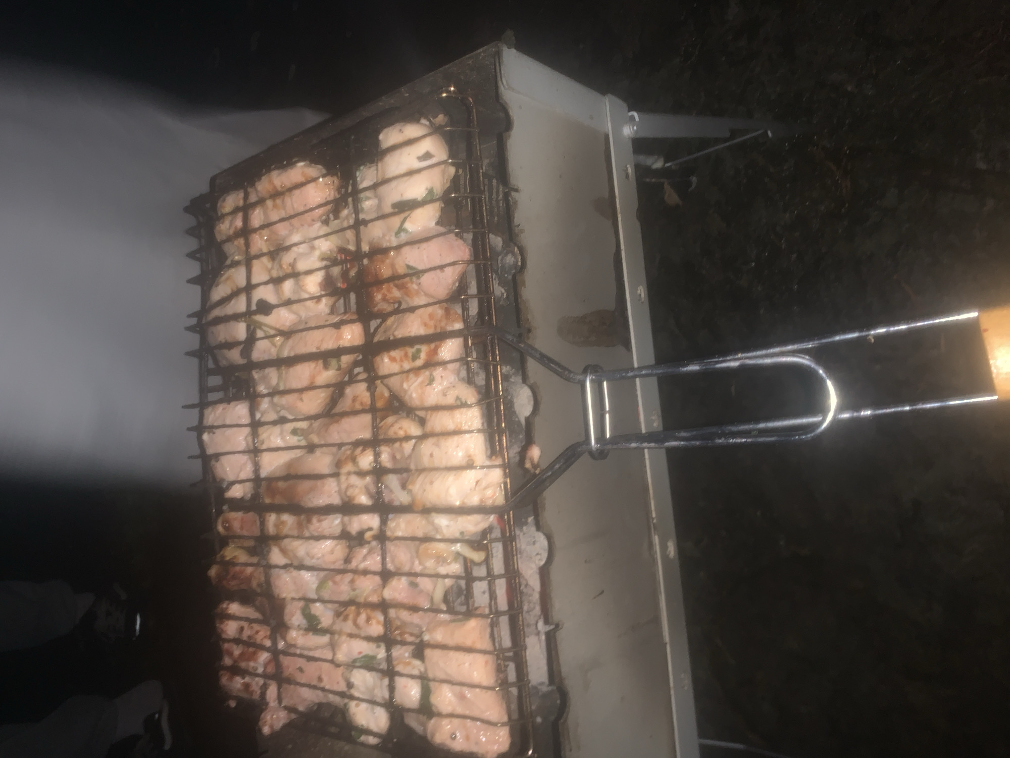 The scene depicts an outdoor nighttime cooking activity in Boitsovo, Russia. A portable, rectangular, light-colored metal barbecue grill is positioned in the lower central and right areas of the frame, resting on visible short legs. A metal grill basket, filled with numerous pieces of raw or partially cooked meat (consistent with chicken or pork), is placed over the grill's heat source. The meat shows evidence of marination, with visible green herbs and small white pieces, possibly onion. Below the grill basket, faint light suggests glowing embers or charcoal, and drips of liquid are visible on the grill's interior. A metallic handle with a wooden grip extends from the grill basket towards the foreground. In the upper left background, the lower legs and feet of two individuals are visible, suggesting their proximity to the grill. One person wears light-colored trousers and dark shoes, while the other wears darker trousers and athletic shoes. The immediate ground and background are dark and indistinct, consistent with an unlit outdoor environment at night.