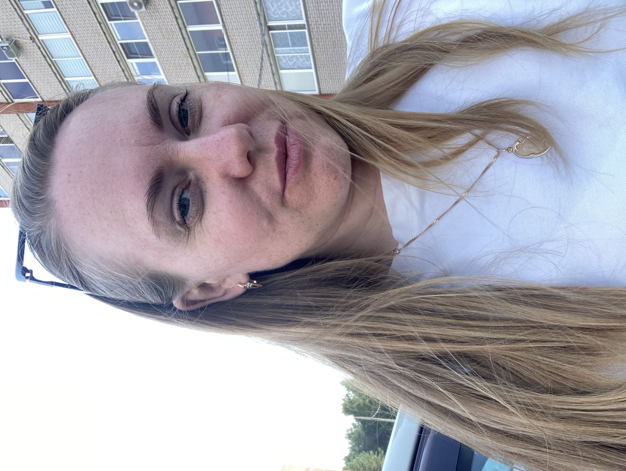 A fair-skinned woman with long blonde hair and freckles is taking a selfie outdoors in front of a multi-story brick building. She is wearing a white t-shirt and a gold necklace with a pendant. She has a gold earring in her left ear and sunglasses perched on her head. The background includes a few trees and a portion of a car on the left side of the frame. The building in the background appears to be an apartment complex, typical of urban areas in Chaikovsky, Russia. The lighting suggests it is daytime, possibly late afternoon, given the angle of the shadows.