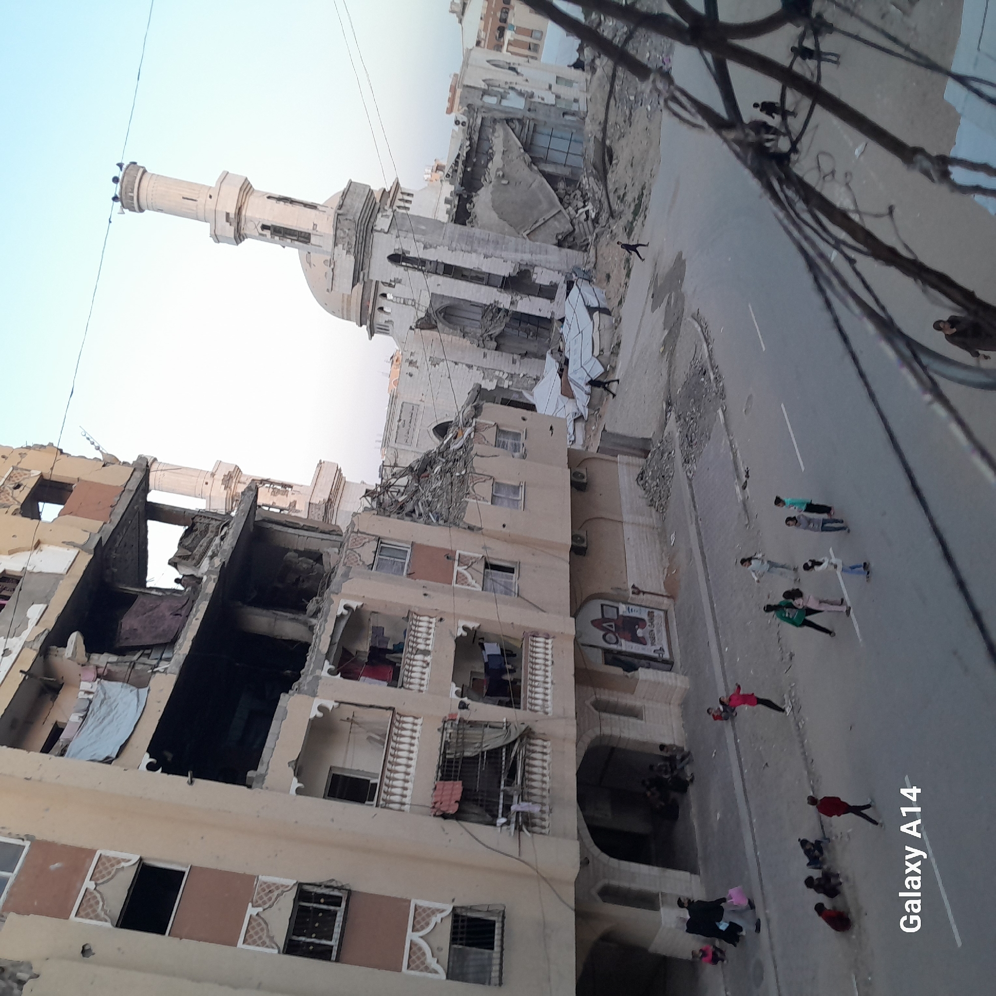 The image depicts a war-torn city street in Unknown, Unknown. A partially destroyed multi-story building stands to the left, its interior exposed with hanging textiles. A mosque with a minaret, also damaged, is visible in the background. A group of people are gathered on the street in the foreground. Electrical wires and poles are present. The scene conveys destruction and potential displacement of civilian population.