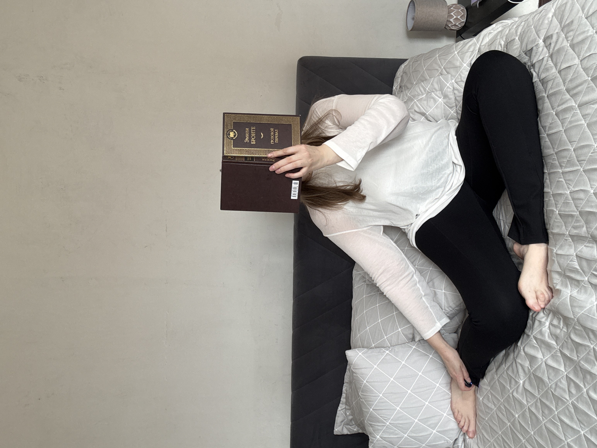 A woman lies on a bed covered with a grey, diamond-patterned comforter. She wears a sheer, long-sleeved white shirt and black leggings. A book titled "Emily Bronte: Grozovoi Pereval" is balanced on her face/head; the book's spine shows the author and title in Cyrillic script. One hand holds the book while the other rests on the bed near her feet. A decorative pillow with a geometric pattern sits at the foot of the bed. The bed has a dark grey headboard. A light grey wall is visible to the left of the bed and a small table lamp with a patterned shade can be seen in the upper right corner of the frame. The image orientation is rotated approximately 90 degrees counter-clockwise. The image's location is unknown, thus I am unable to confirm it is in Izhevsk, Russia.