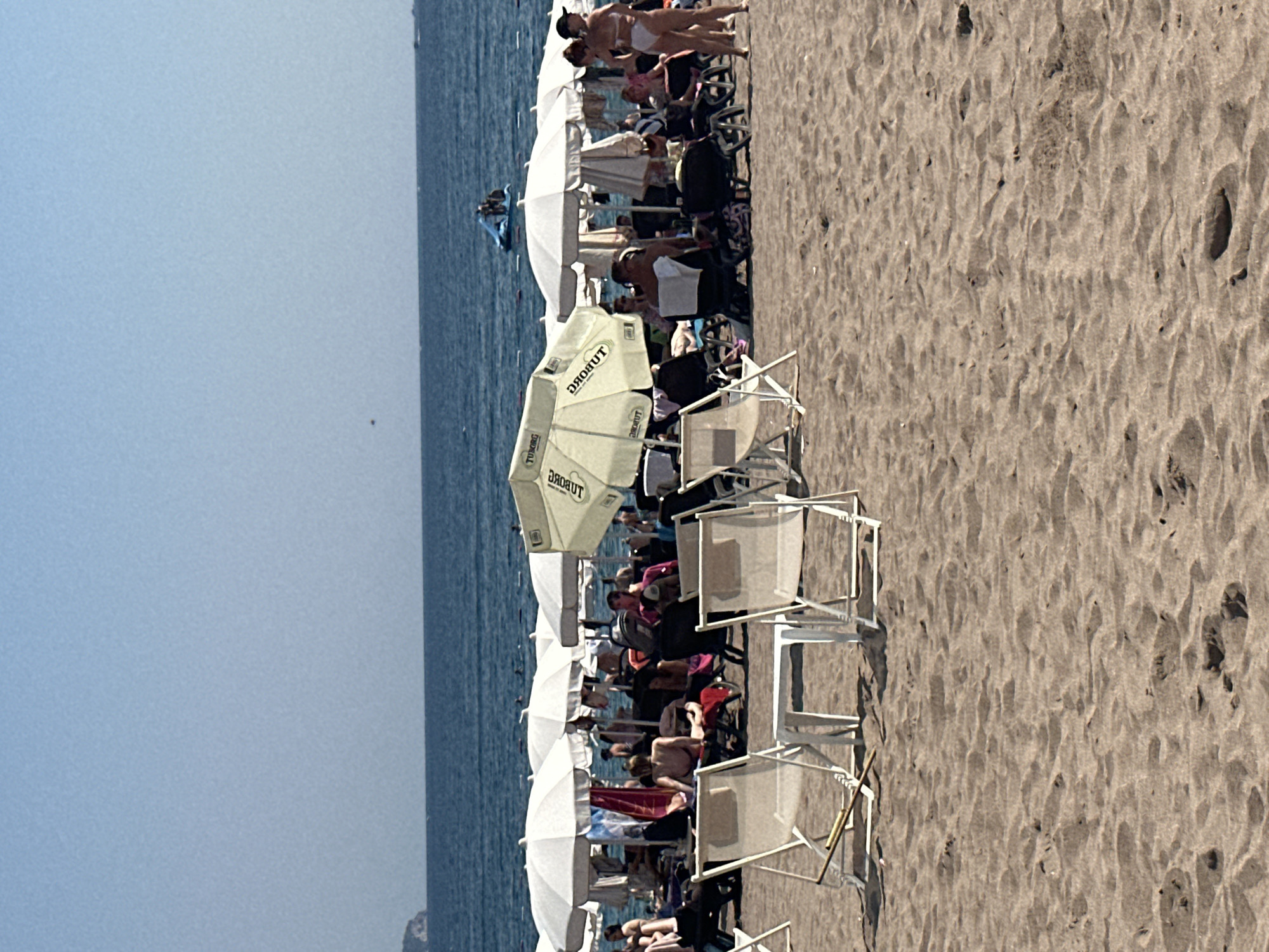 The image depicts a crowded beach scene in Boreti, Montenegro, on a sunny day. The foreground is dominated by sand with footprints and two empty white beach chairs and a small white table. Behind them, numerous white beach umbrellas and loungers are set up, with many people relaxing on them or walking around. A few individuals are visible in the background, some in swimwear. The sea is visible beyond the beach, with a clear blue sky above. In the middle distance, on the water, there's a blue inflatable water slide with people on it. The overall atmosphere is one of a popular tourist destination enjoying recreational activities.