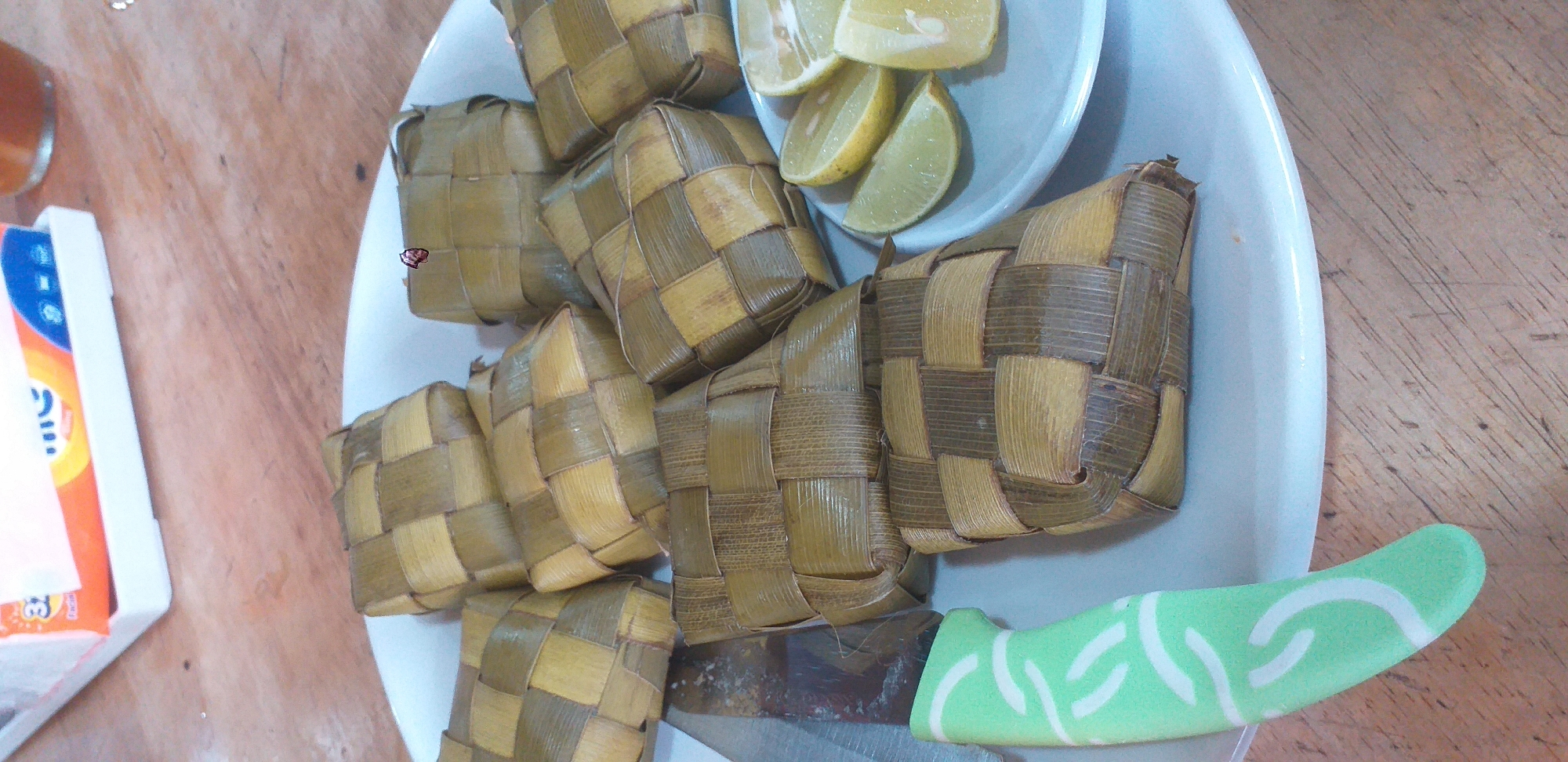 A top-down shot captures a white plate piled with Ketupat, traditional Indonesian rice cakes, and lemon wedges in Batulicin, Indonesia. A knife with a green and white handle rests on the plate, near the Ketupat. A tissue box is visible in the background, partially obscured. The scene takes place on a wooden surface.