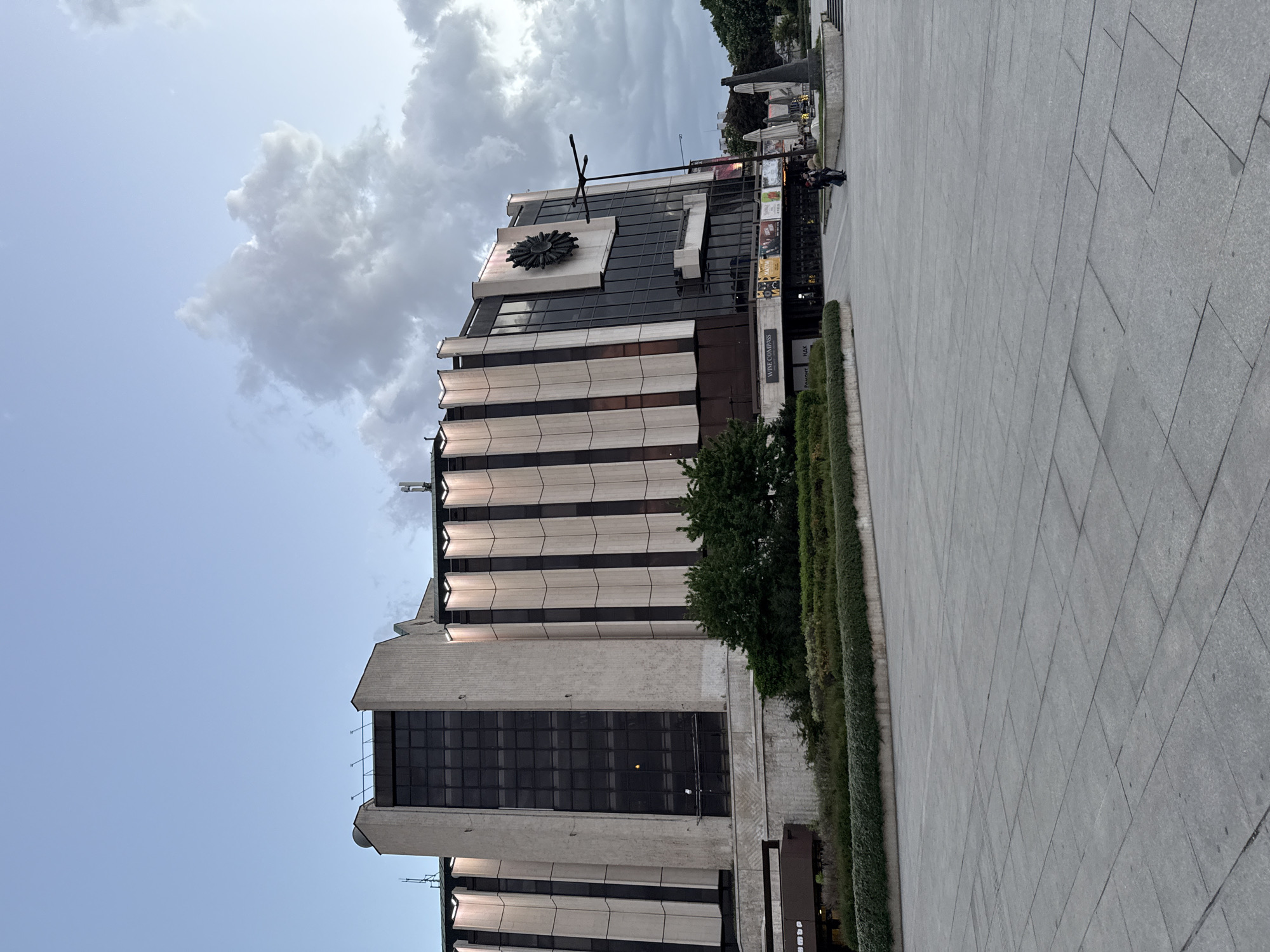 The image captures the National Palace of Culture (NDK) in Sofia, Bulgaria. The NDK is a large, brutalist-style building with a striped facade of alternating white and dark bands. A prominent ornamental sun-like symbol is visible on the upper section of the building. Below, there is a sign for "Wine Compass". A well-maintained hedgerow borders the building. The foreground shows a paved public square made of large gray tiles. In the background, there are some individuals seated in an outdoor area, possibly a cafe or seating area. The sky above is a mix of blue and gray, with a large cloud formation.