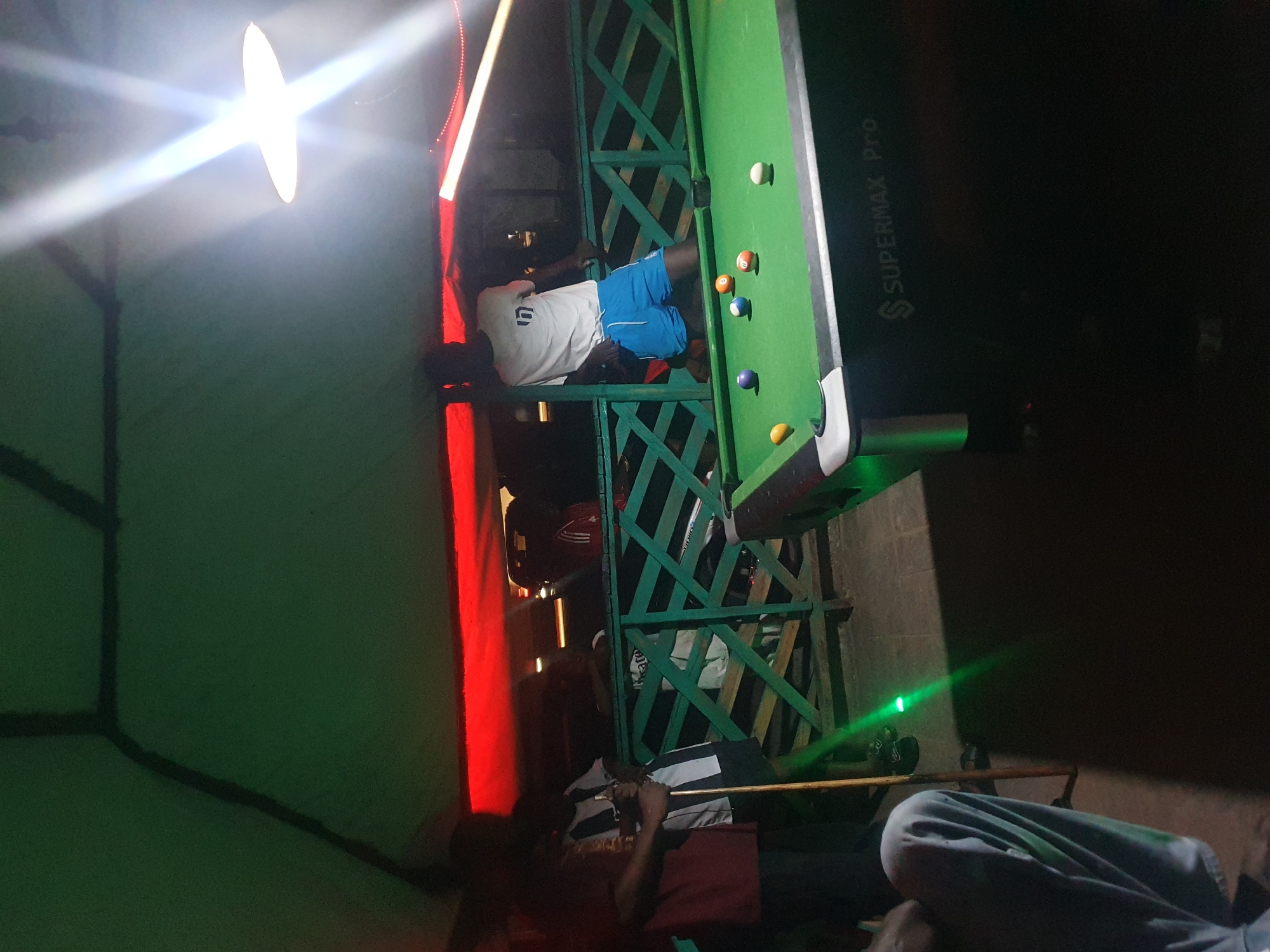 In Lokoja, Nigeria, at a dimly lit outdoor establishment with a green overhead covering and red lighting accents, a group of individuals are gathered around a billiard table. The scene is illuminated by bright overhead lights, casting dramatic shadows. On the green felt of the billiard table, several balls are scattered, including a yellow cue ball, a purple ball, a blue ball, and an orange ball with the number '11' visible. To the right of the table, a person wearing blue shorts and a white t-shirt with the number '7' is leaning against a green, diamond-patterned railing. A person in a maroon shirt and a black and white striped shirt is standing near the table, holding a pool cue. Another individual in a light blue garment is seated in the foreground, partially obscured. The backdrop features wooden structures and vehicles, suggesting a recreational or social gathering space. The billiard table itself is labeled "SUPERMAX Pro". The atmosphere appears casual and social.