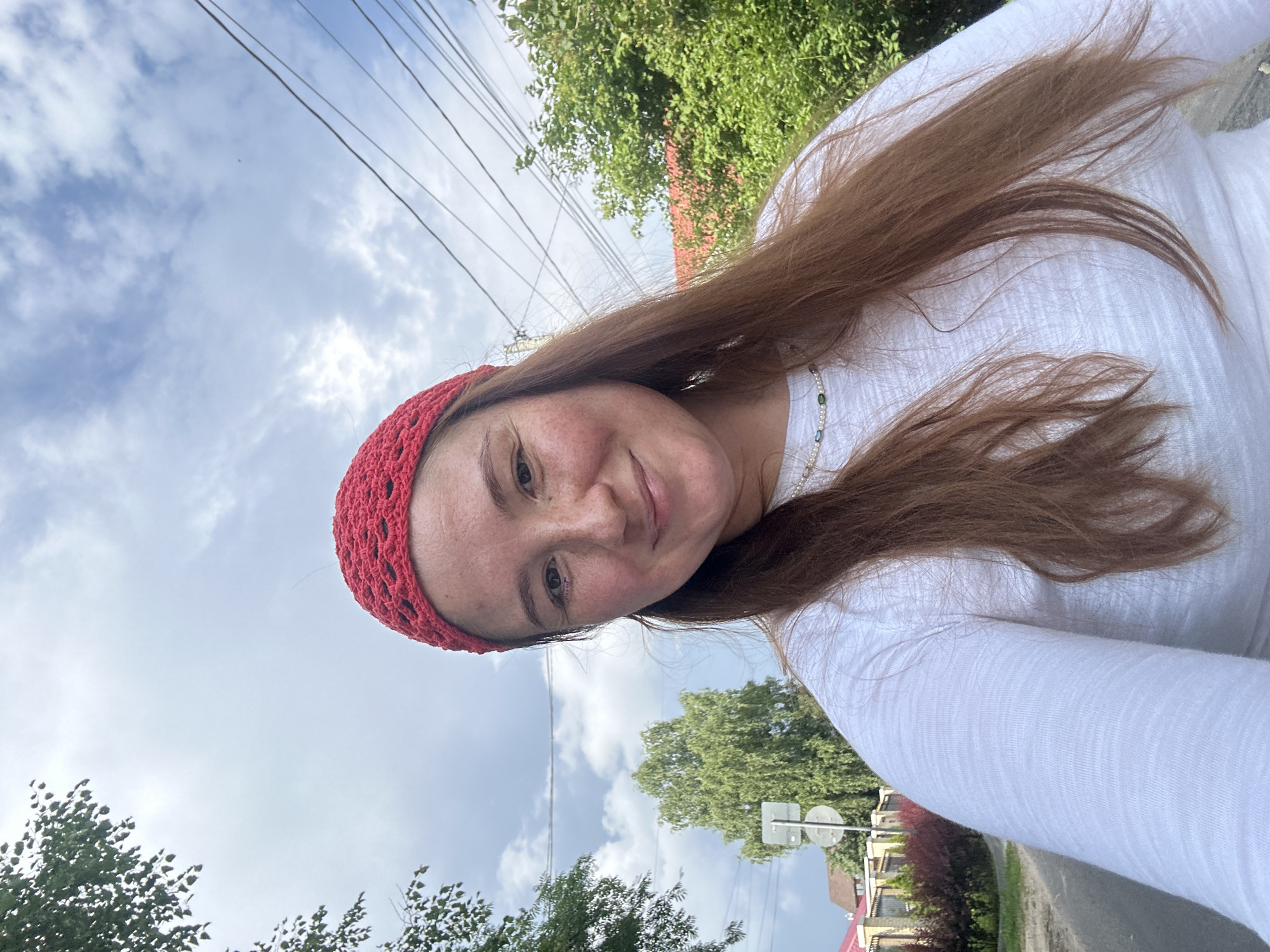 In Nizhny Novgorod, Russia, a young woman with long, reddish-brown hair and a red crocheted headband is captured in a selfie. She wears a white long-sleeved shirt and a delicate necklace. Her face is turned towards the camera, and she has a light smile. Freckles are visible across her nose and cheeks, and subtle makeup is applied to her eyes. The background reveals a cloudy sky with visible power lines overhead. To the left, lush green trees frame the scene, and a street sign is partially visible. Further back, a fence and houses suggest a residential area. The overall environment appears to be an outdoor setting in a city, possibly a street or park.